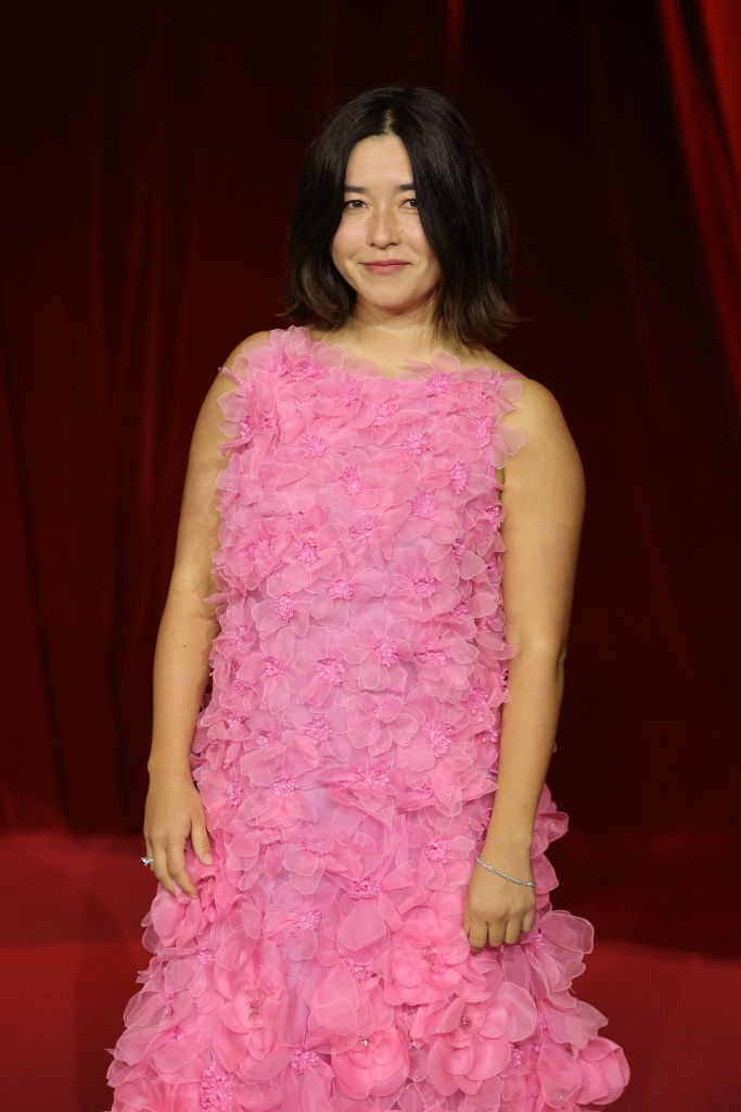 A woman in a floral-textured, sleeveless dress stands on a red carpet