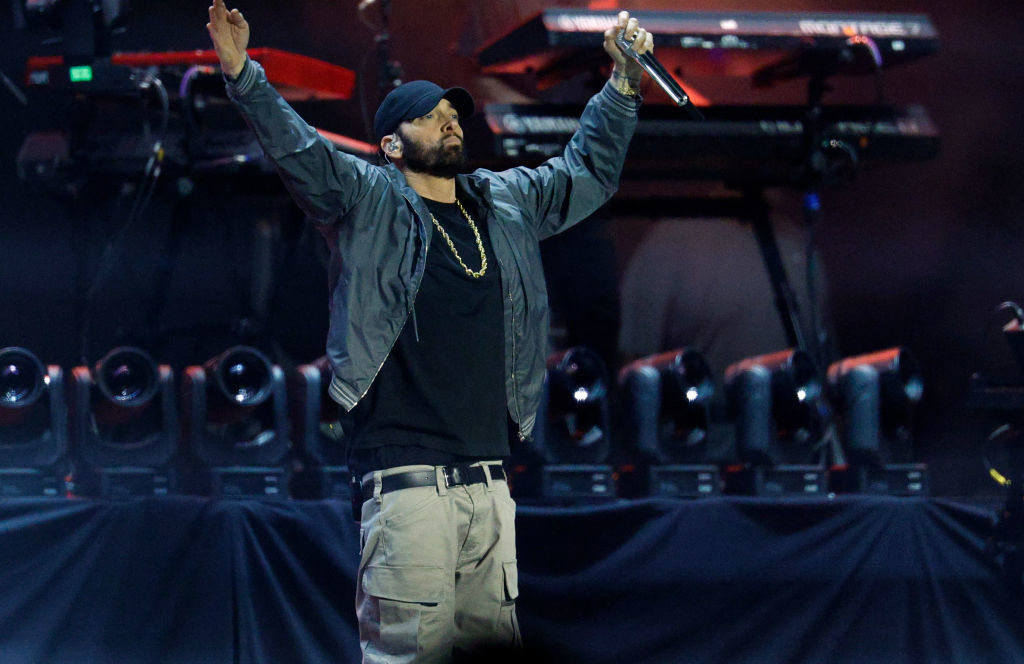 Eminem on stage wearing a jacket and cargo pants, holding a microphone, with hands raised during a live performance