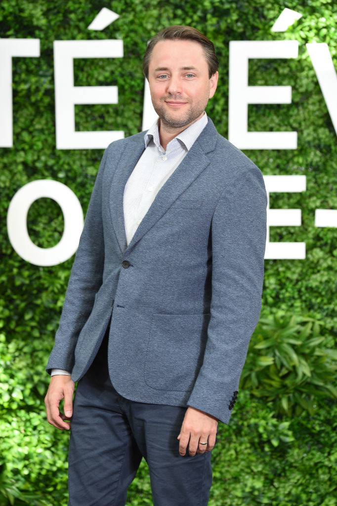 Vincent in a suit jacket and white shirt smiles in front of a greenery wall at a celebrity event