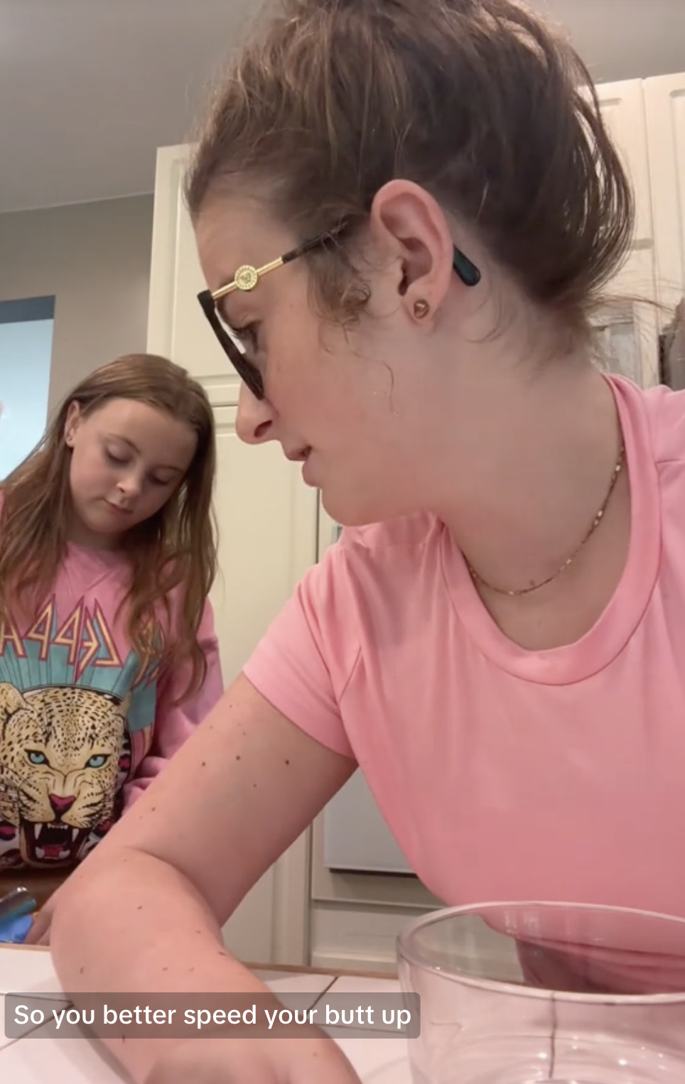 Ali wearing eyeglasses leans over a table while Addie in a graphic sweatshirt stands nearby. Caption reads, &quot;So you better speed your butt up&quot;