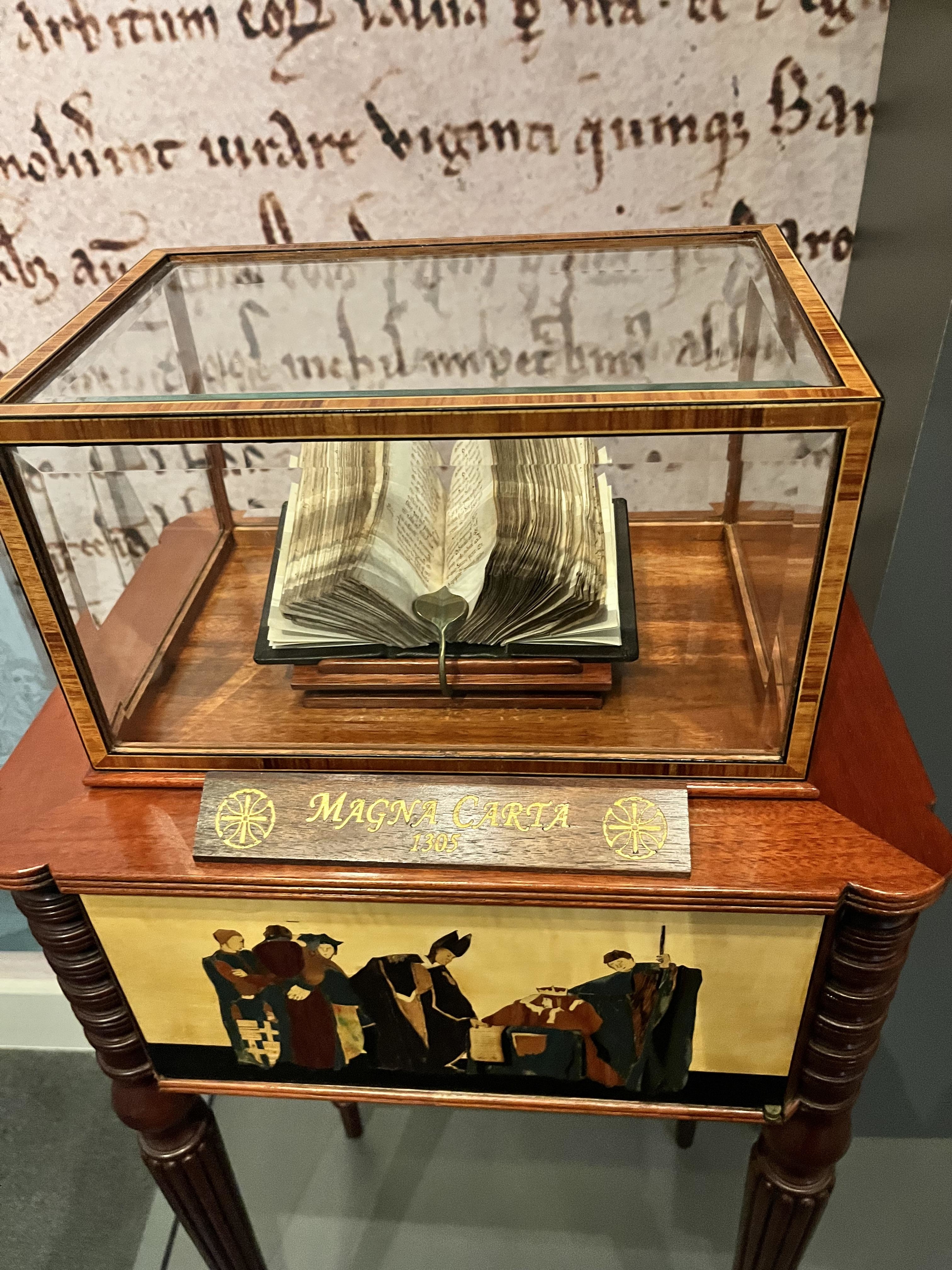 The Magna Carta displayed in a glass case on a wooden stand with historical text in the background