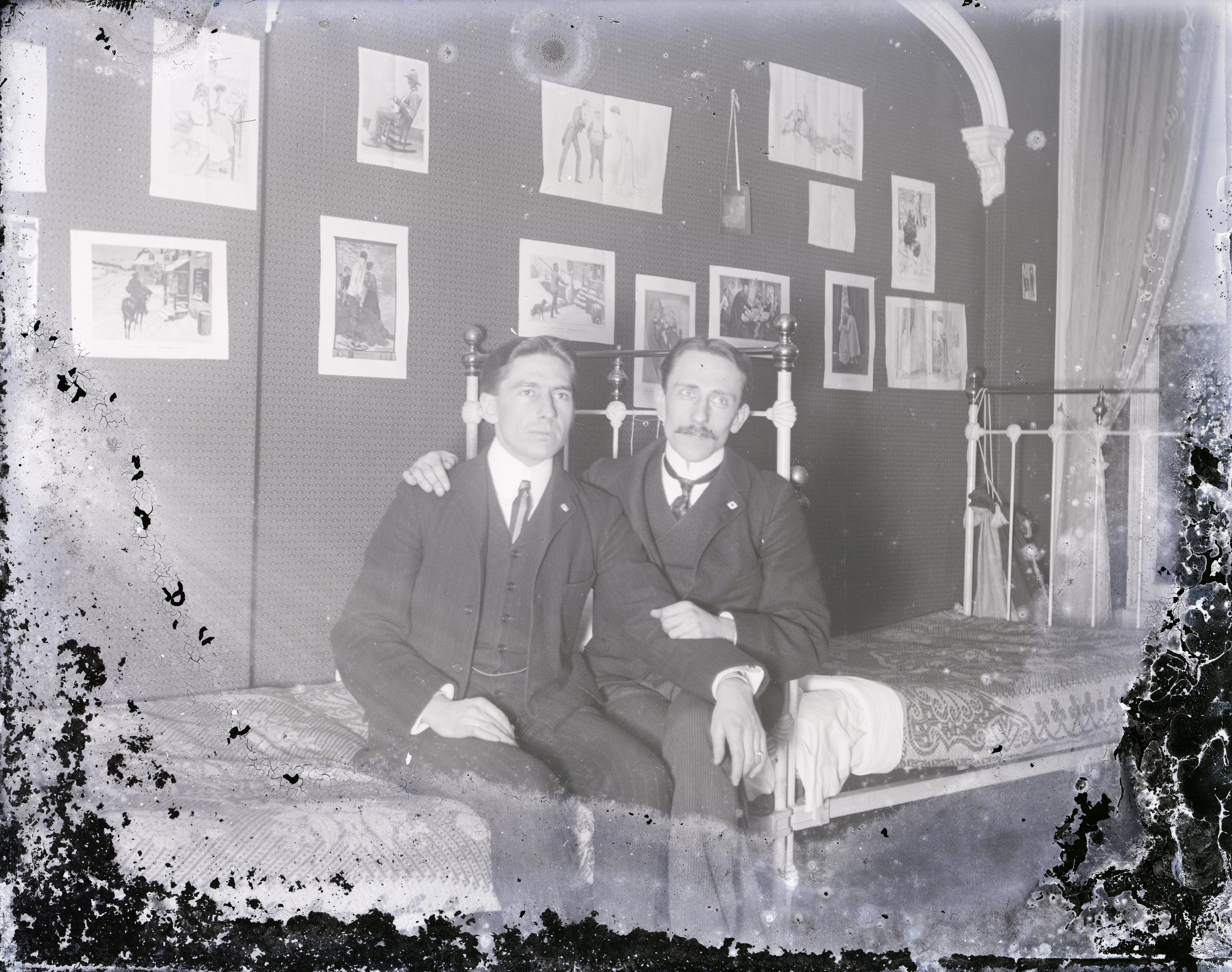 Two men seated on a bed in a decorated room with framed pictures. They are wearing formal suits