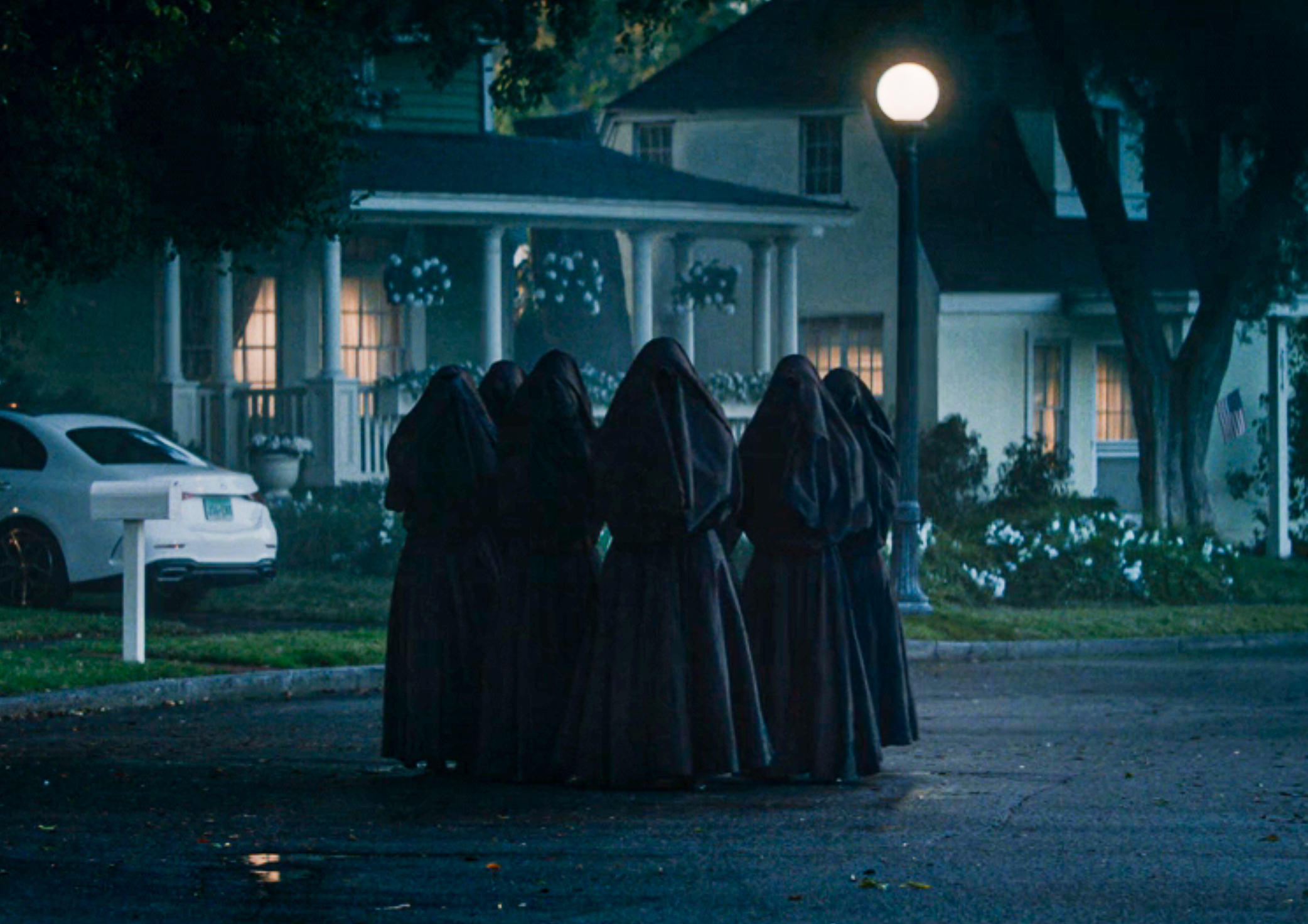 A group of people in dark cloaks stands in a circle on a quiet street at night, near a house with lights on