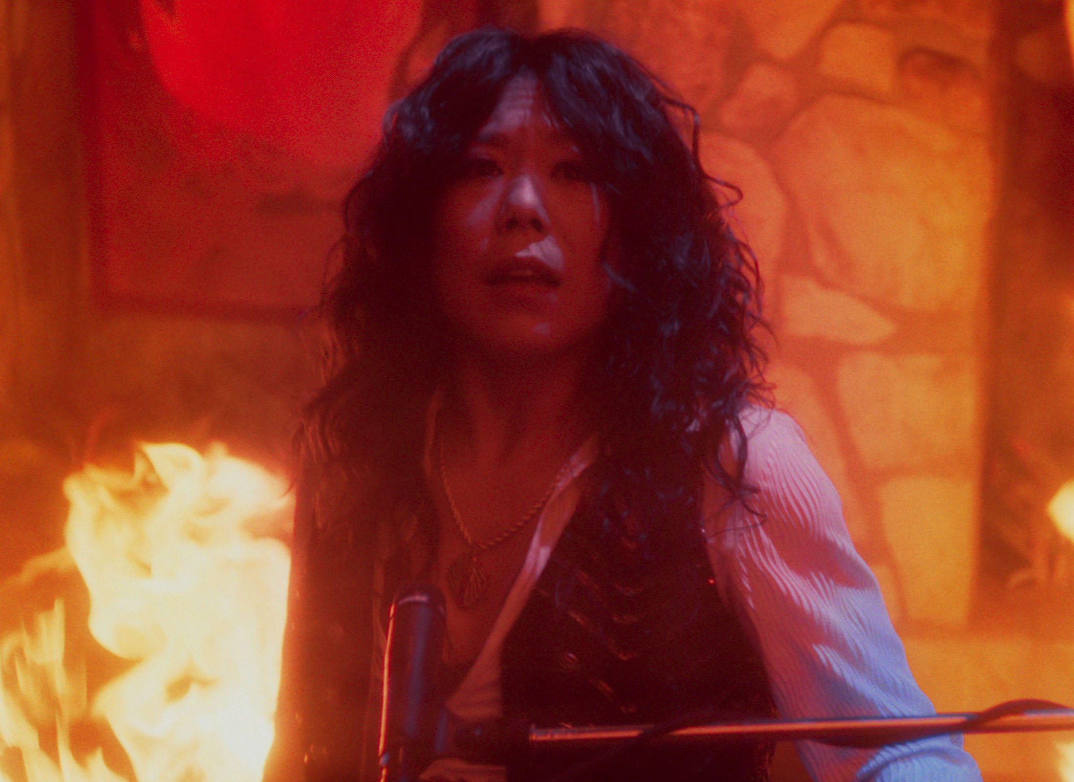 A person with long, curly hair performs passionately on stage, surrounded by flames and music equipment