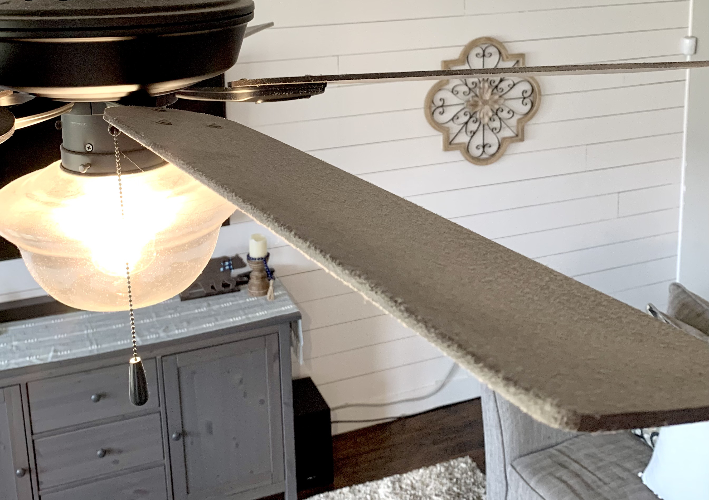 Close-up view of a dusty ceiling fan blade in a cozy living room with wooden furniture and wall decor