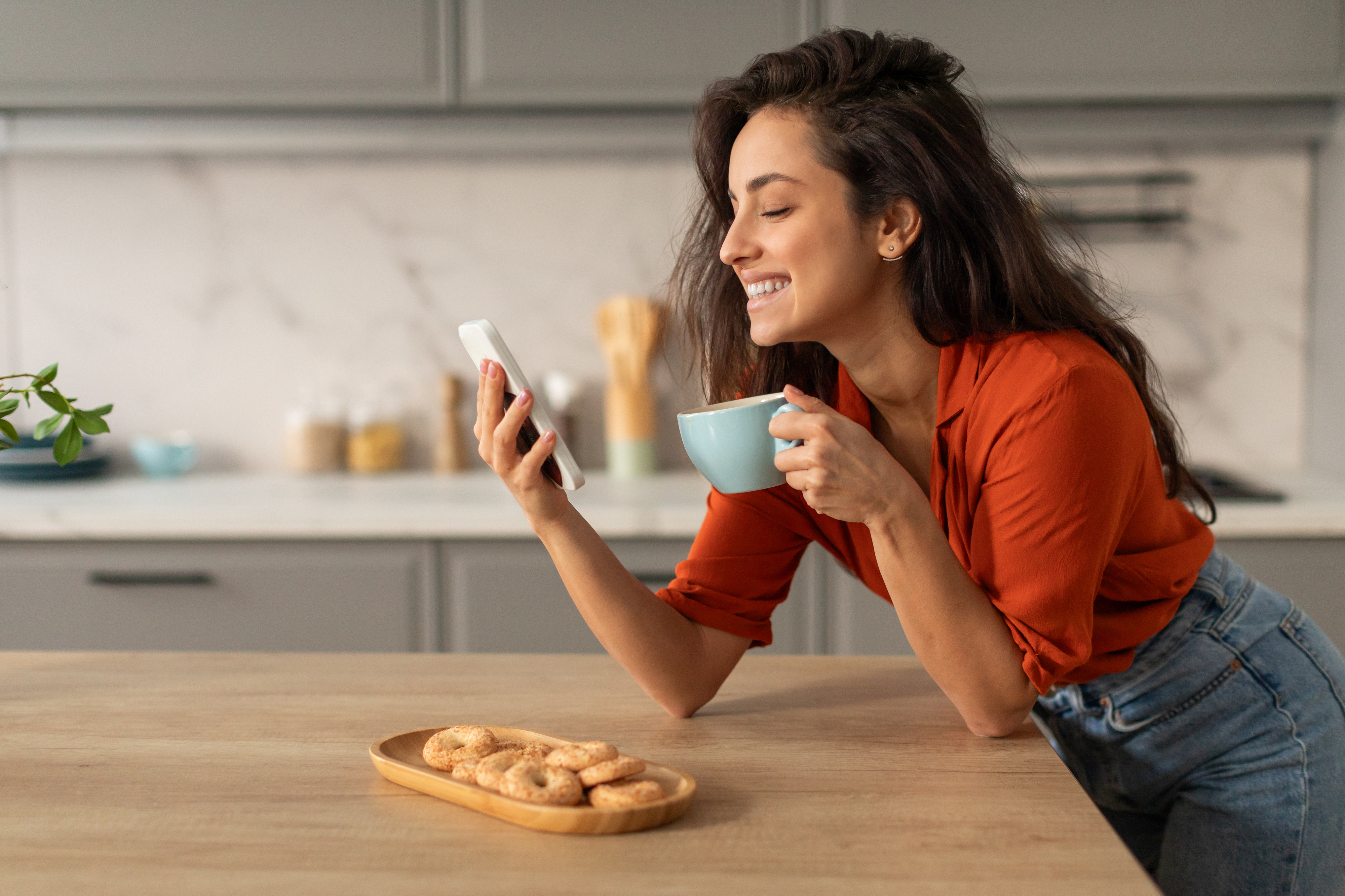 A person holding a phone and a cup, smiling in a kitchen with cookies on the counter