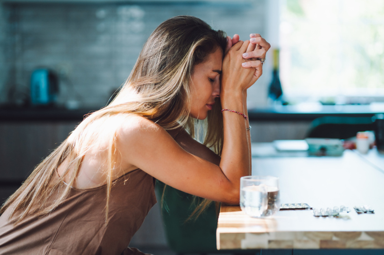 A woman sits at a table with her head on her hands, appearing thoughtful or stressed, next to a glass of water and medication strips