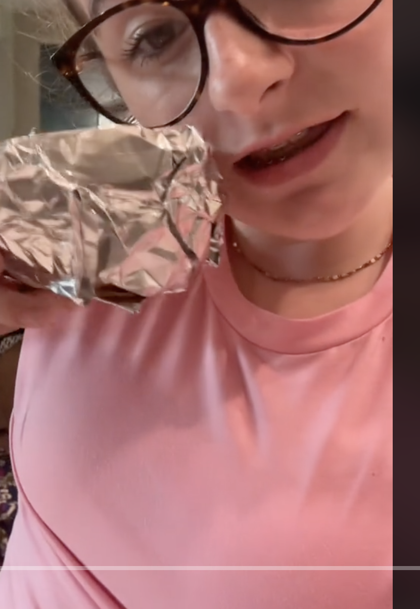 Addie holding a foil-wrapped object close to their face, appearing to gesture or explain something