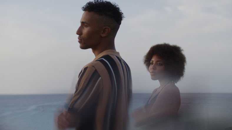 A man and woman stand by the ocean, looking into the distance with thoughtful expressions