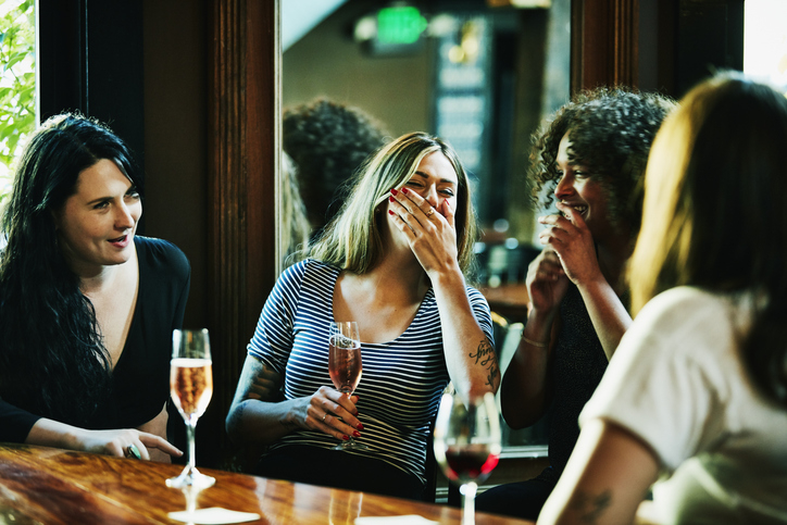 Four people sharing a laugh at a bar, each holding a drink, seated at a wooden table