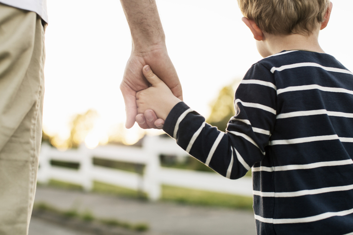 Adult and child holding hands, child wearing striped shirt, background shows a white fence and blurred outdoor scenery