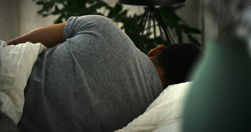 Person sleeping on their side in bed, partially covered by a blanket, room dimly lit