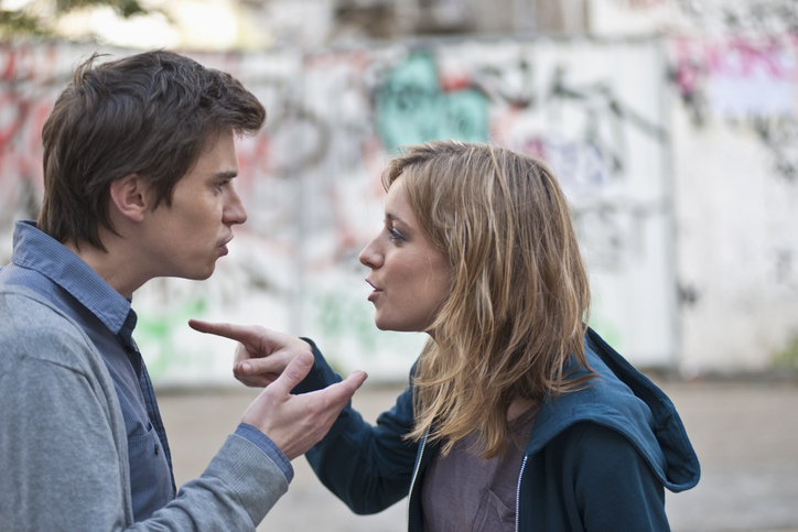 A man and woman are having a heated discussion outdoors, gesturing and facing each other closely with intense expressions