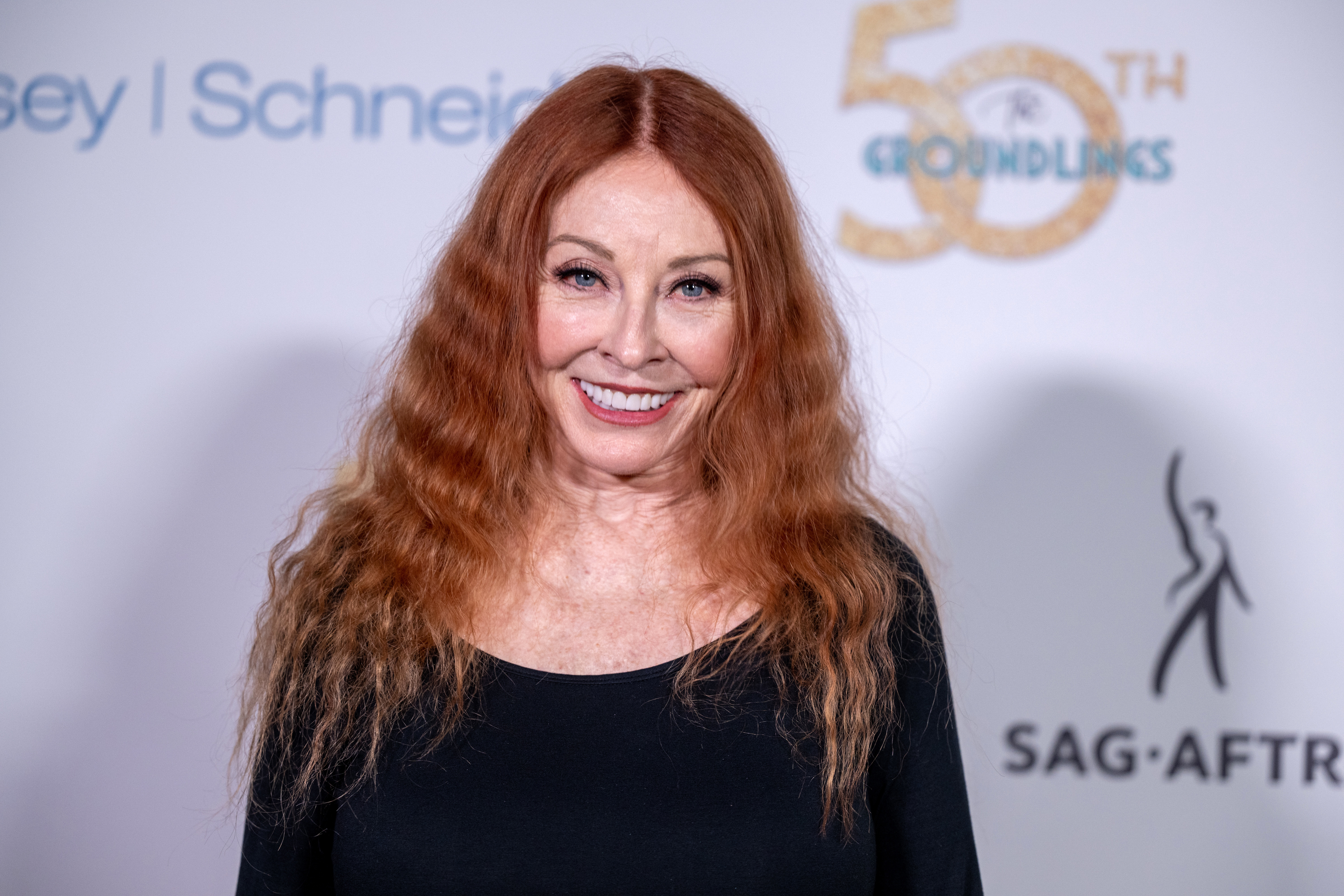 Cassandra with long, wavy hair smiles on a red carpet. She wears a simple top. The backdrop mentions SAG-AFTRA and a 50th event