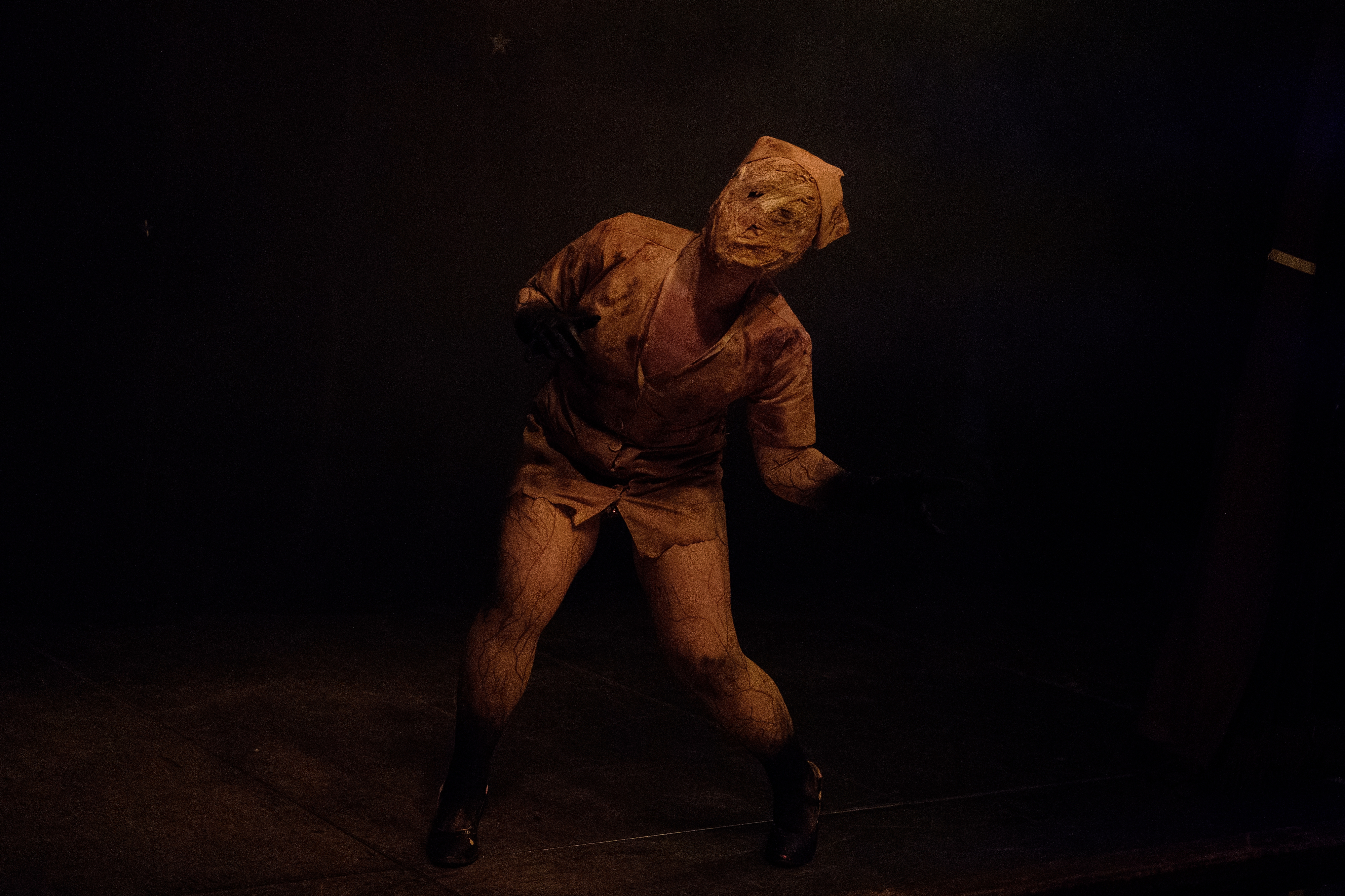 A person in a wrapped mask and distressed outfit poses dramatically on stage, evoking a mysterious or horror theme