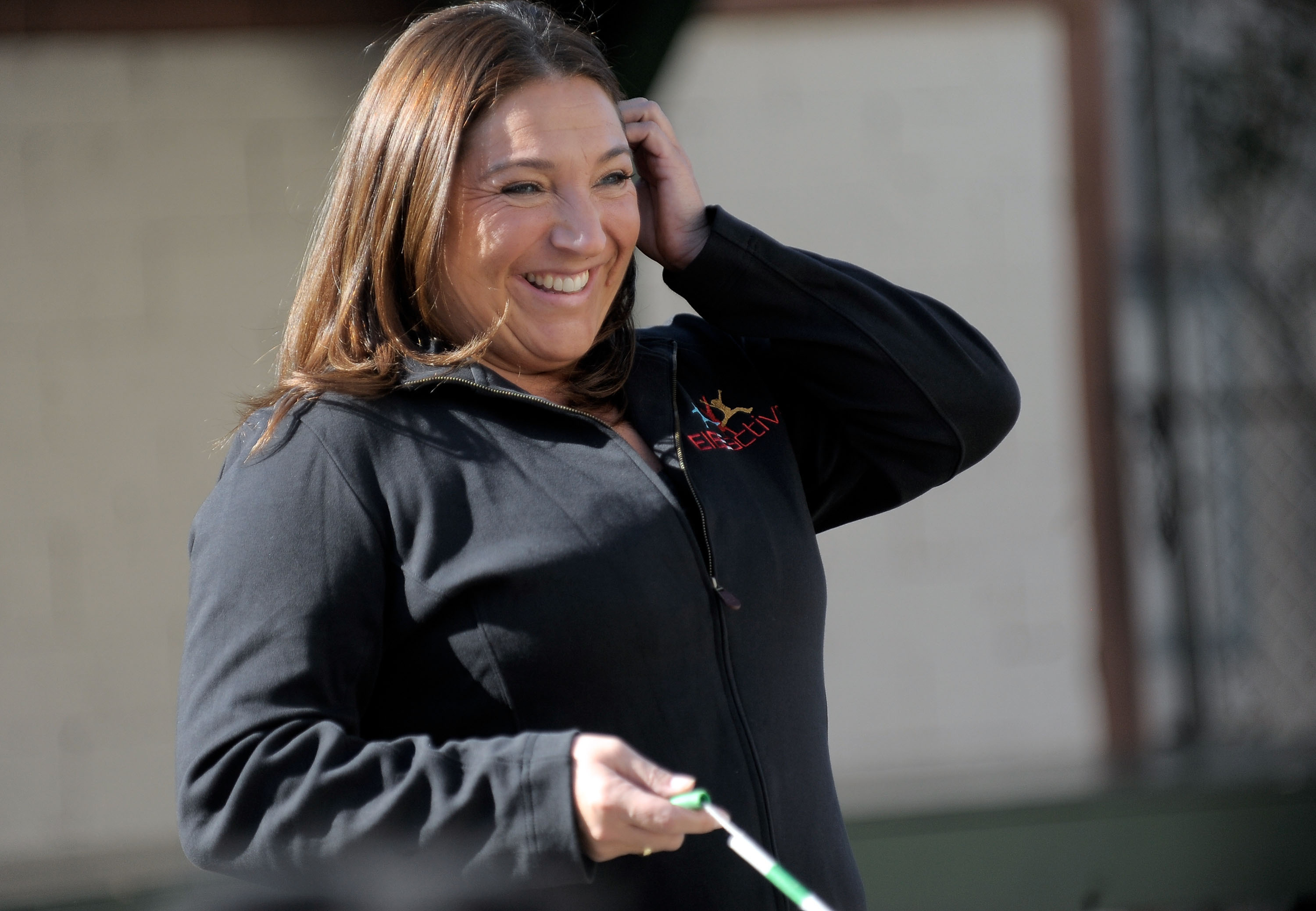 A woman with brown hair smiles, wearing a black jacket, holding a walking stick