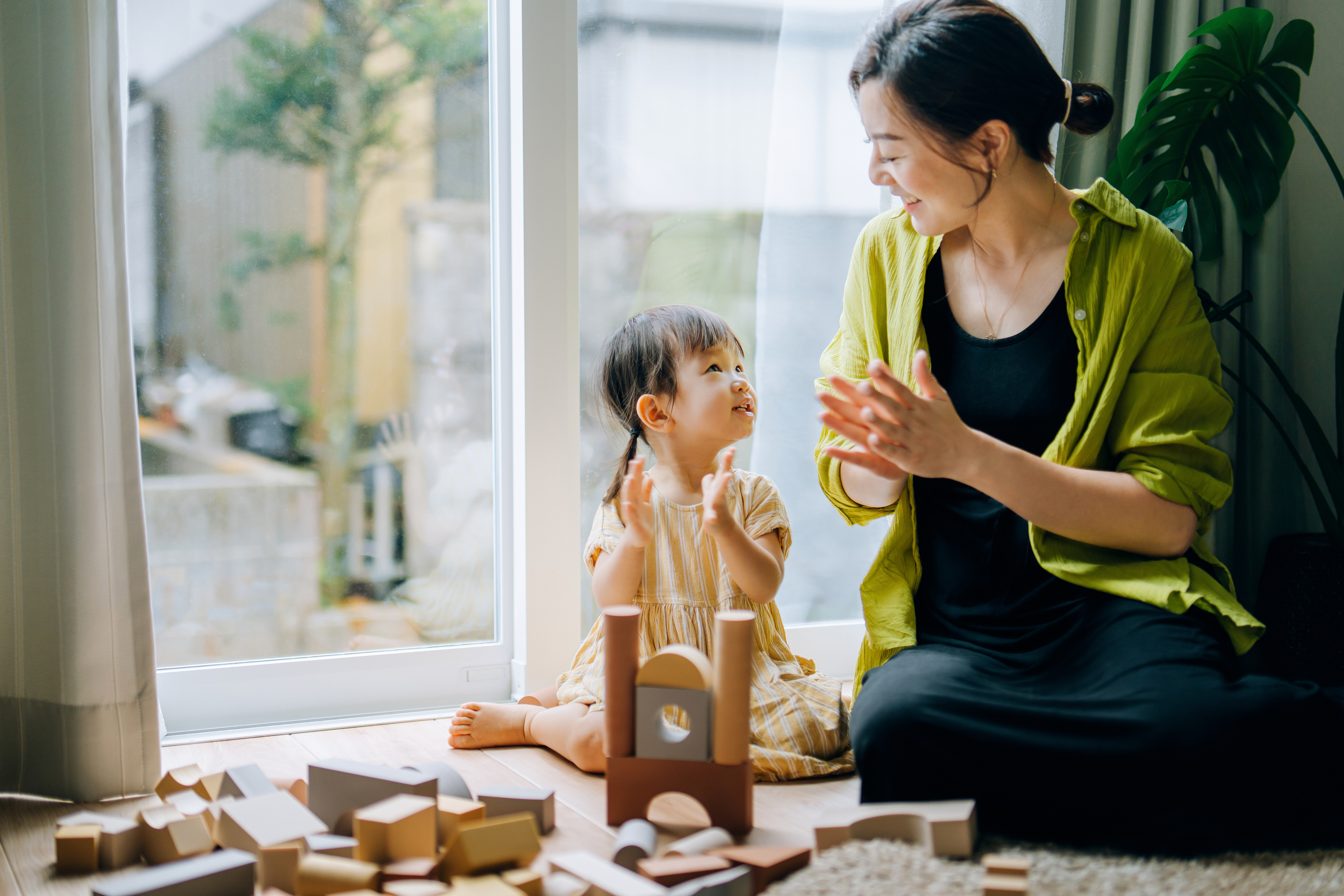 A woman and child sit by a window playing with wooden blocks, smiling at each other