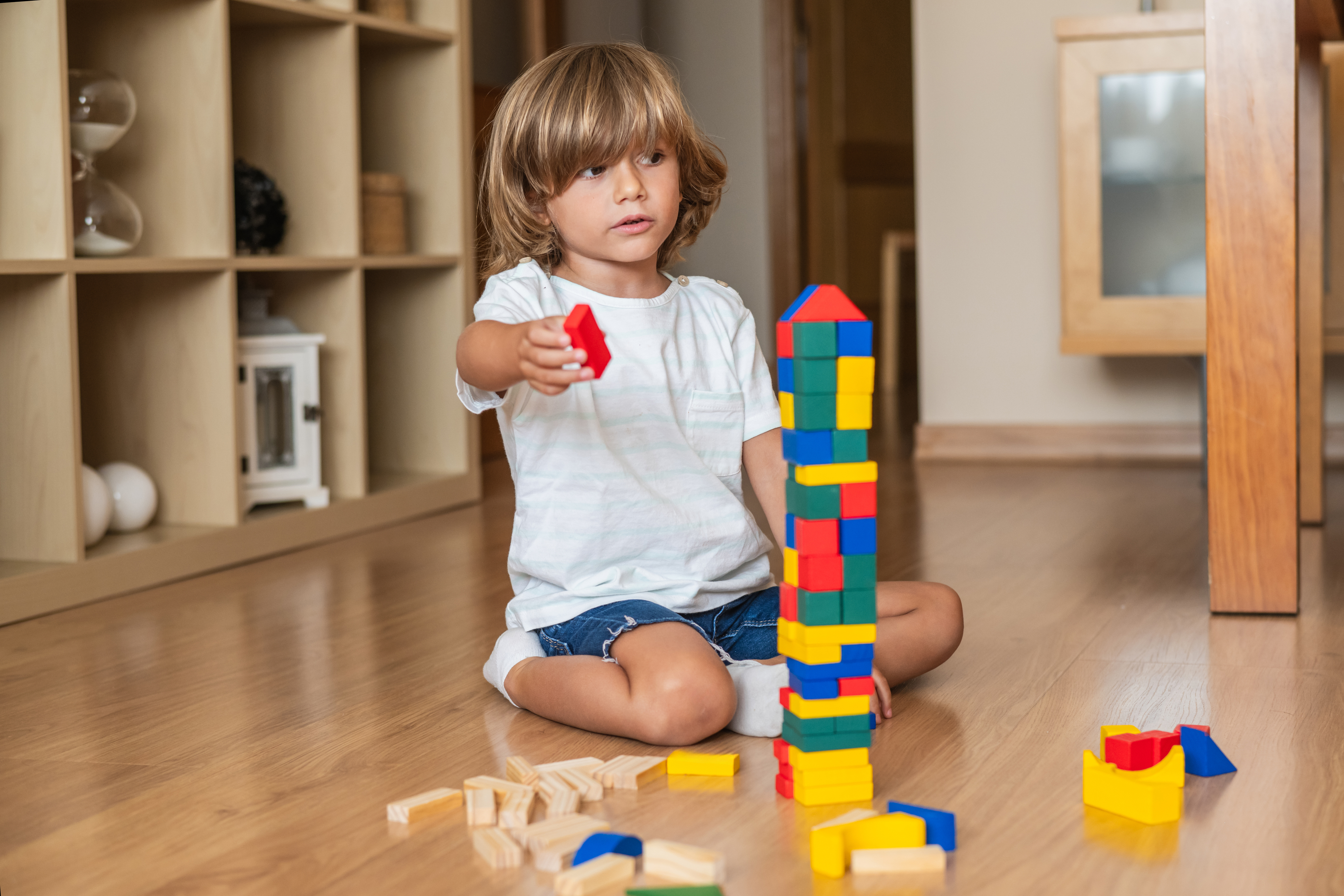 Child plays on the floor, building a tall tower with colorful blocks, concentrating on adding a red block