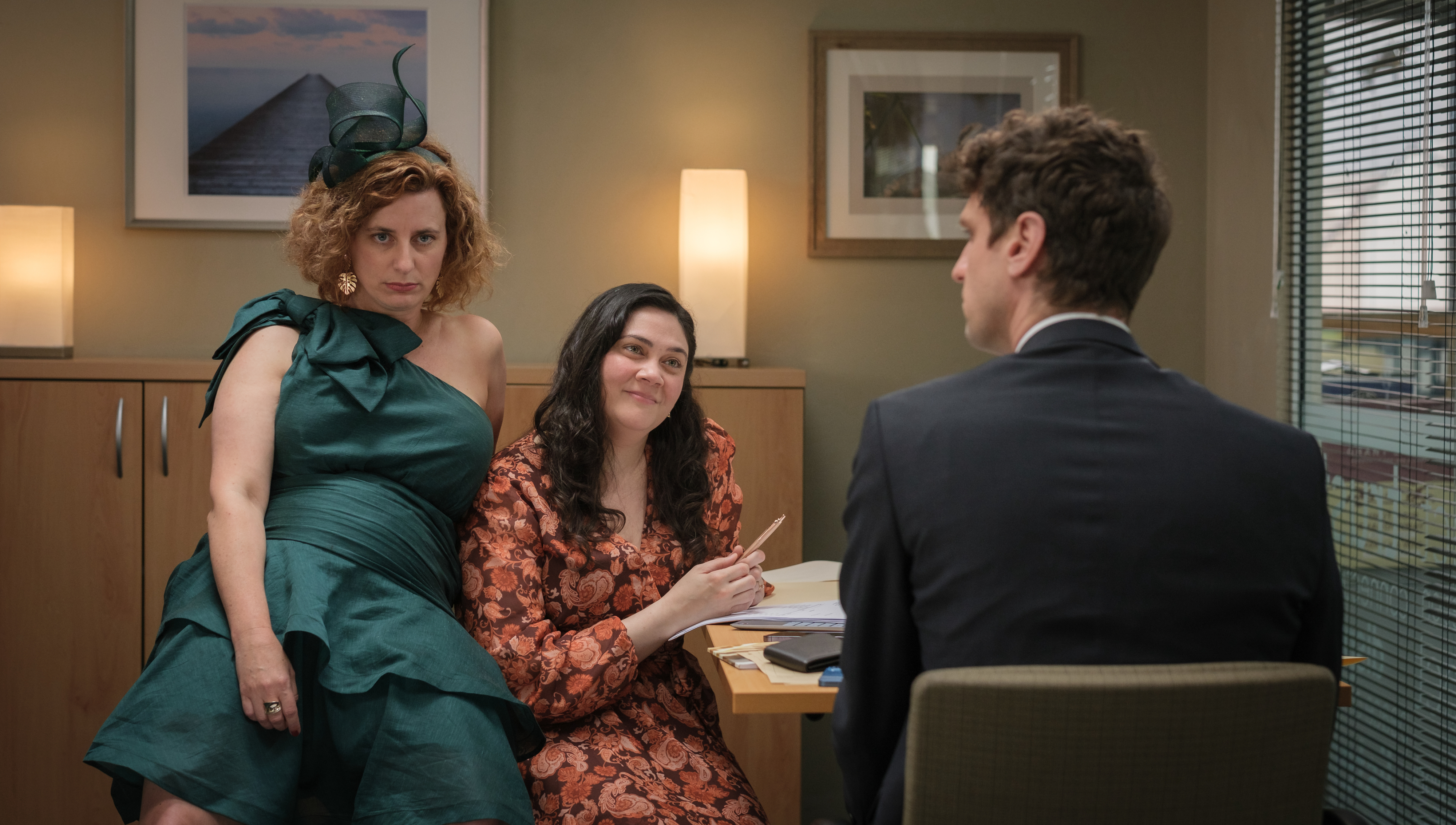 Two people in party dresses sit across from a man in a suit, engaged in conversation in an office setting