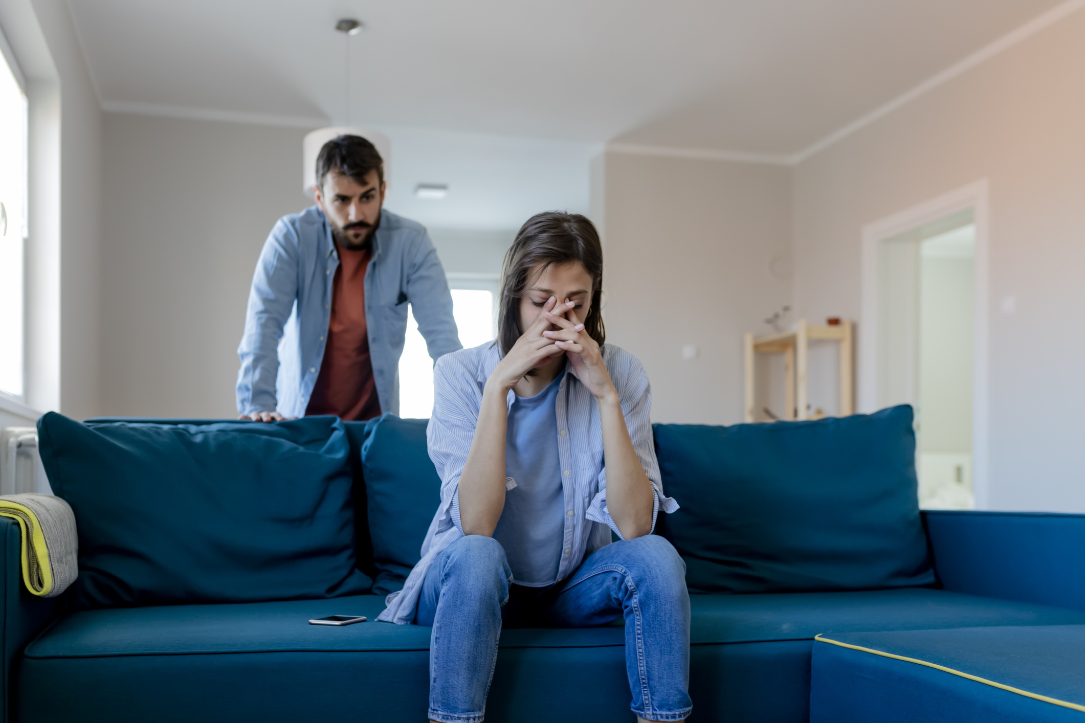 A woman sits on a couch looking distressed, while a man stands behind her appearing concerned