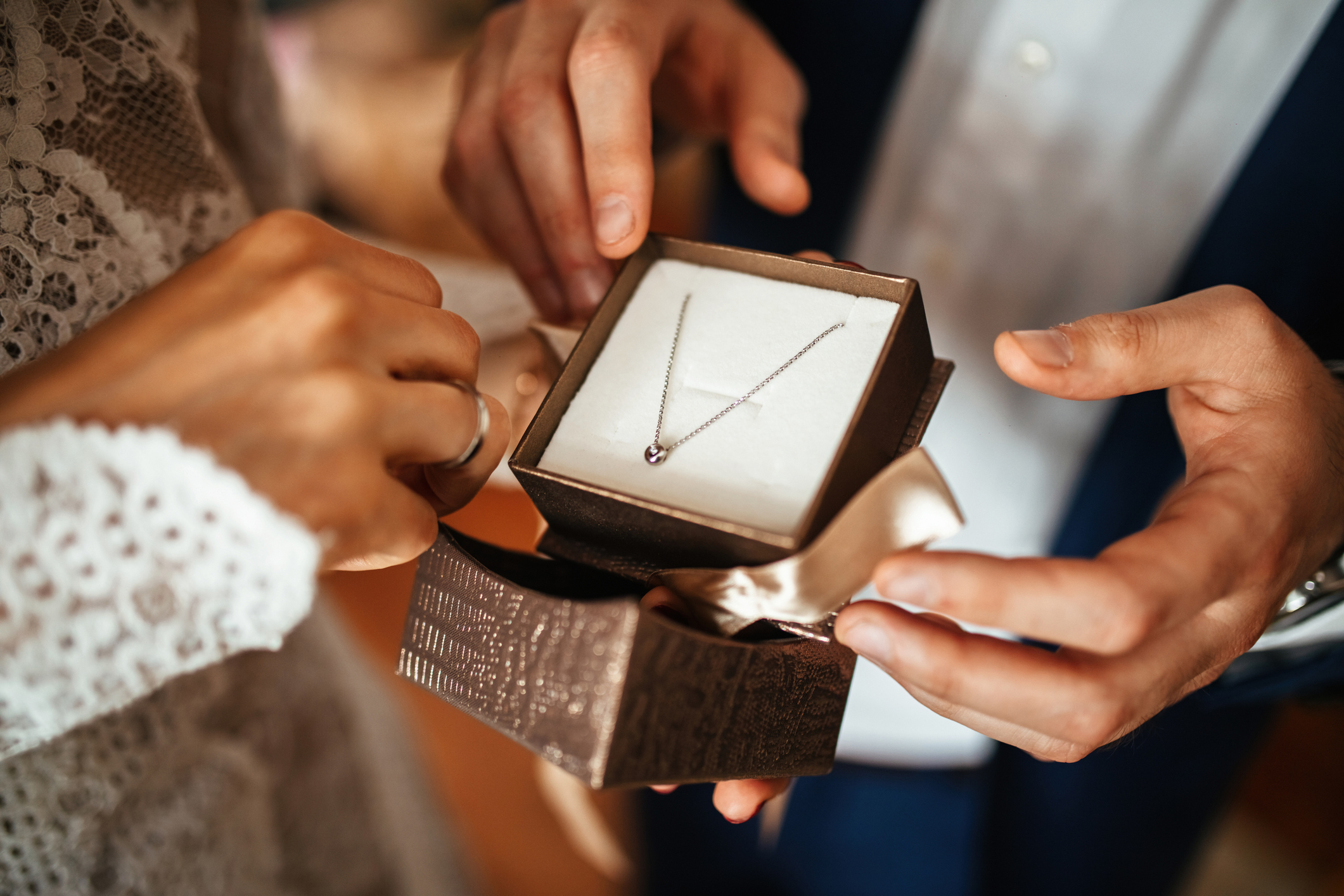 A person presents a jewelry box with a delicate necklace to someone in lace attire, suggestive of a romantic gesture or gift exchange