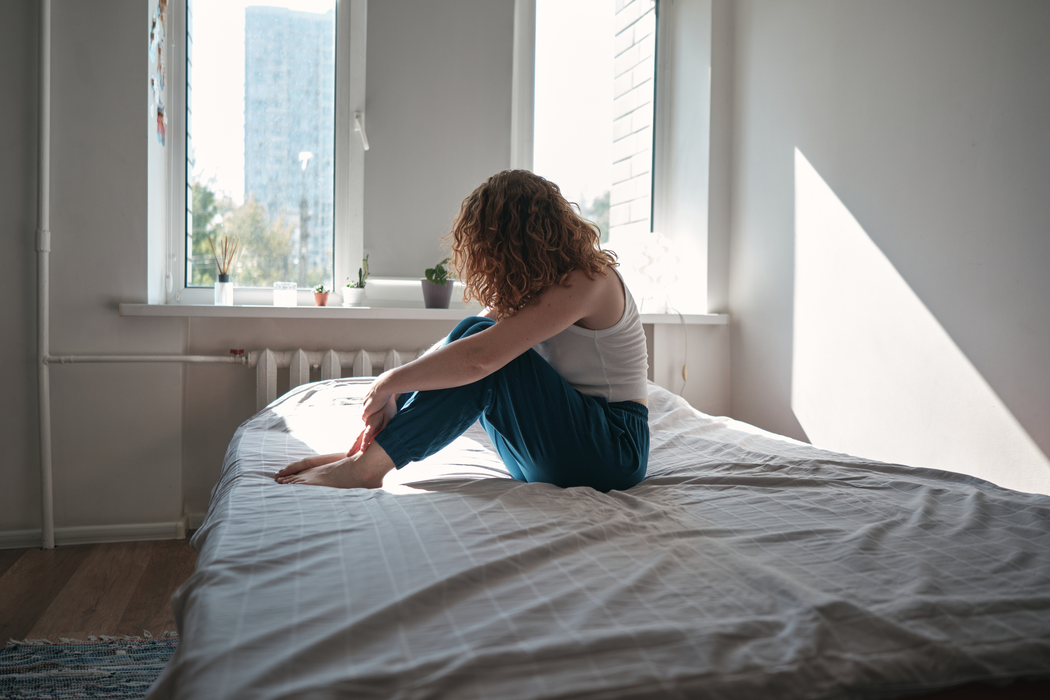 Person sitting alone on a bed, looking out the window. They have curly hair and are wearing casual clothing