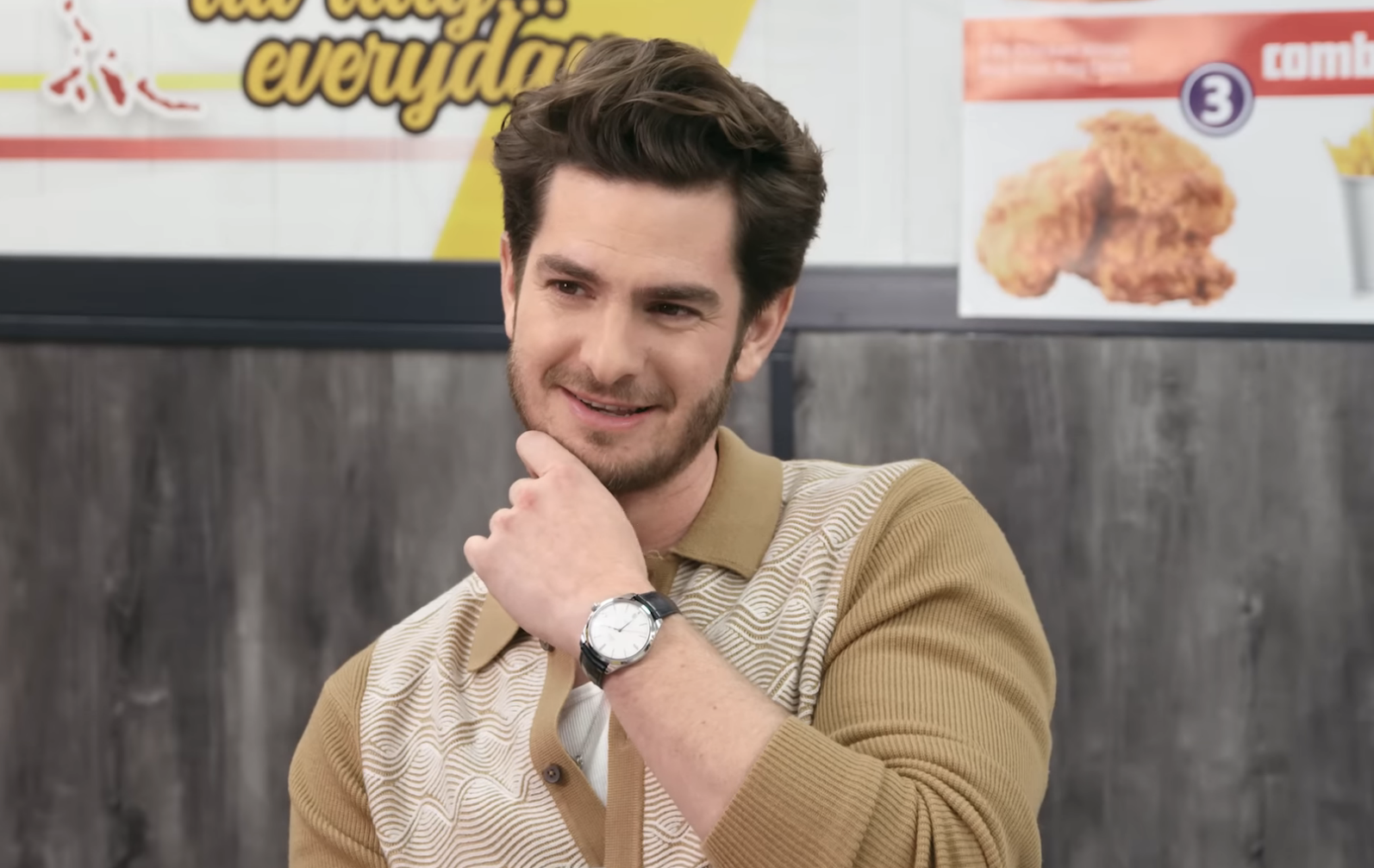 Andrew Garfield smiling in an interview for "Chicken Shop Date" with Amelia Dimoldenberg, seated at a table with food