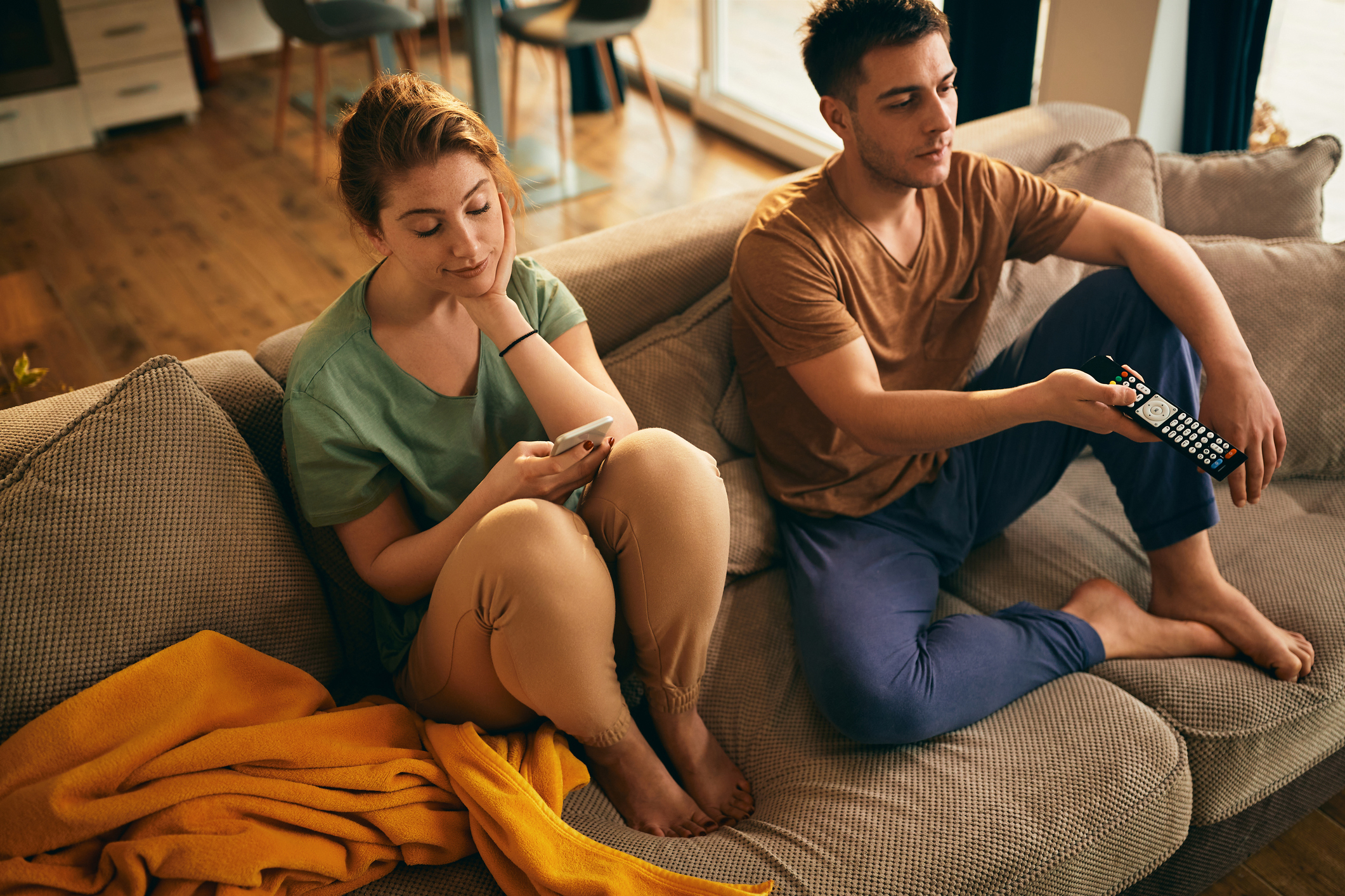 A couple sits apart on a couch; one looks at a phone, the other holds a remote, highlighting disconnection