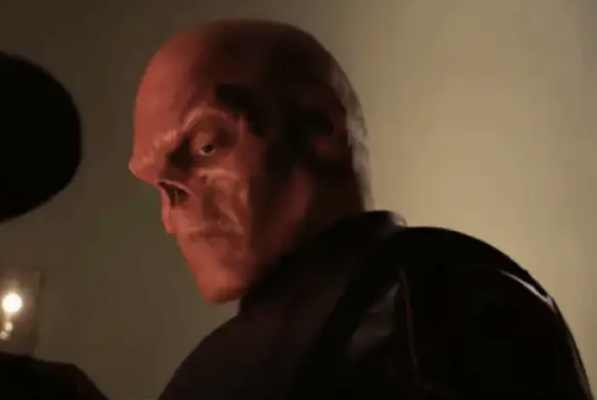 Hugo with a skull-like face, wearing a dark outfit, in a dramatic scene