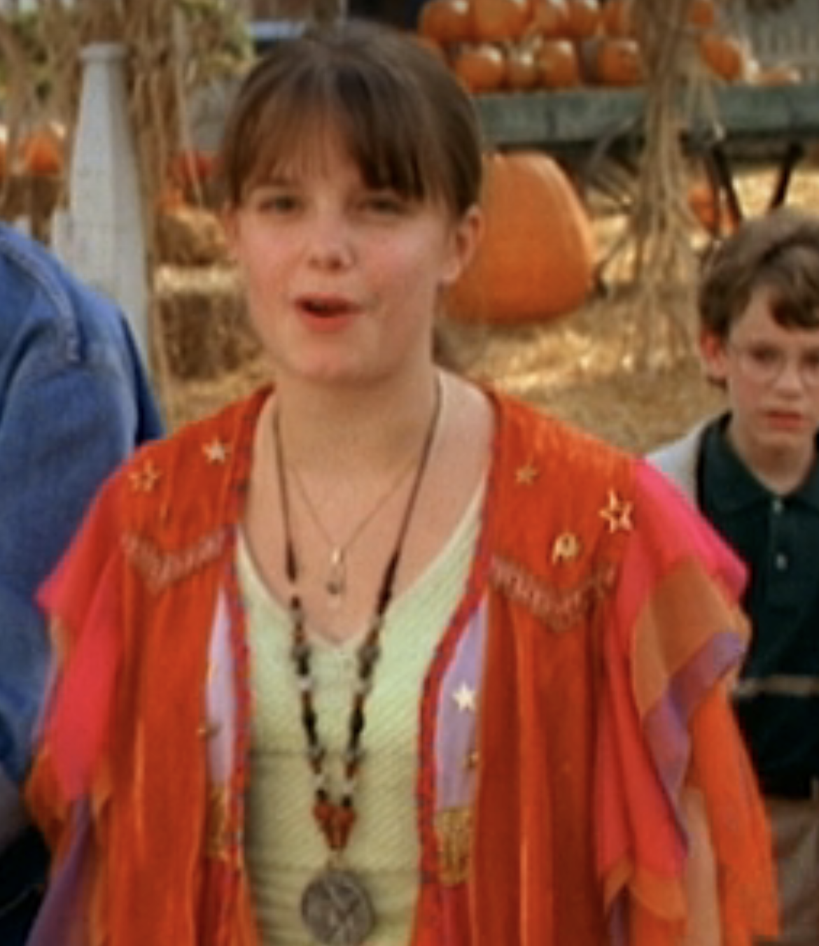 Kimberly, in a flowy shawl and long, beaded pendant necklace over a t-shirt, standing confidently in front of hay bales and pumpkins