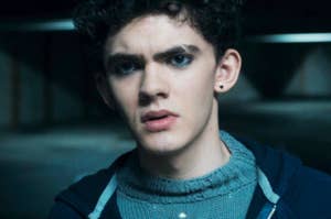 Joe Locke with curly hair and dark eyeliner wearing a knitted sweater under a jacket, looking contemplative in a scene from "Agatha All Along"