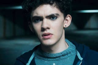 Joe Locke with curly hair and dark eyeliner wearing a knitted sweater under a jacket, looking contemplative in a scene from 