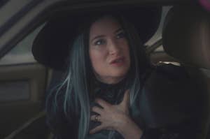 A woman with long dark hair and a large hat sits in a car, looking up with her hand on her chest