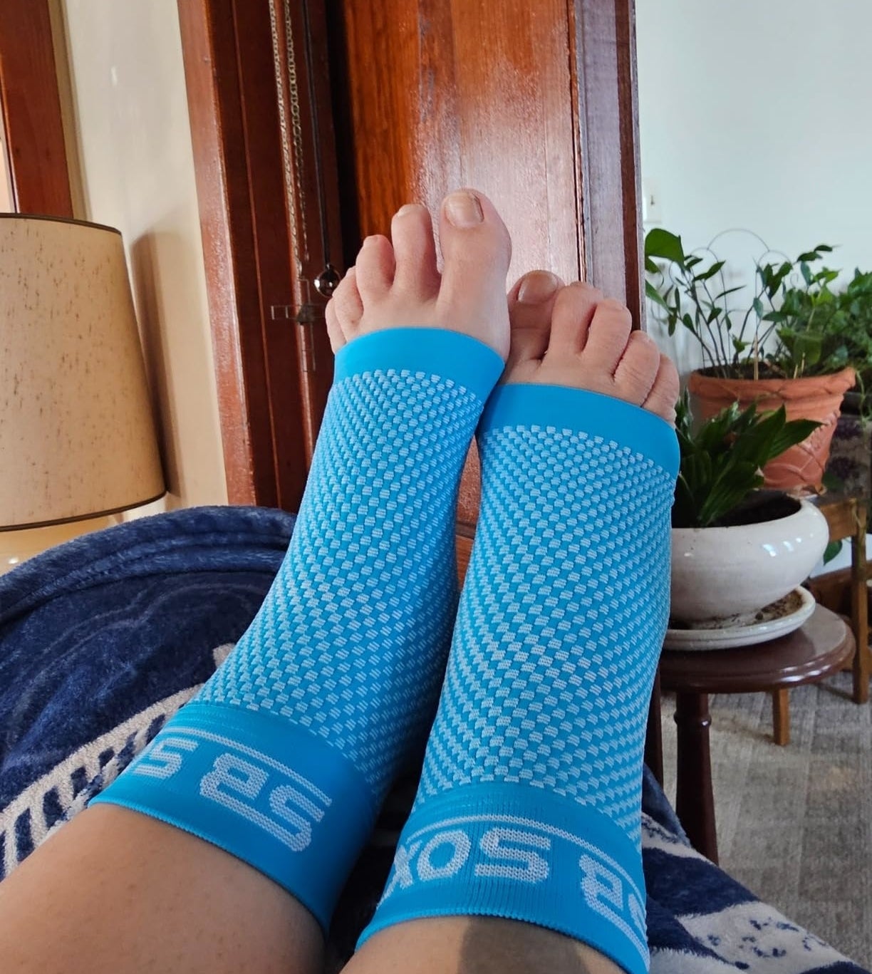 Person wearing blue compression socks with dotted pattern, feet resting on a blanket, indoor setting with plants in background