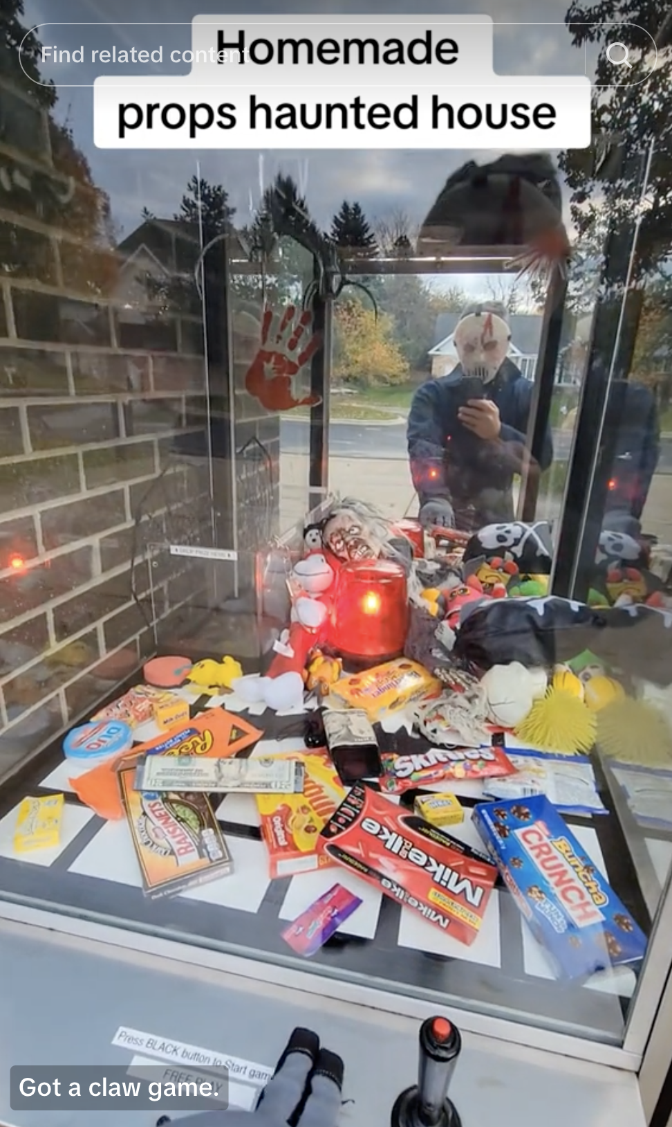 Claw machine filled with Halloween props and candy outside. Person in a mask taking a selfie in the reflection. Text: &quot;Homemade props haunted house&quot;