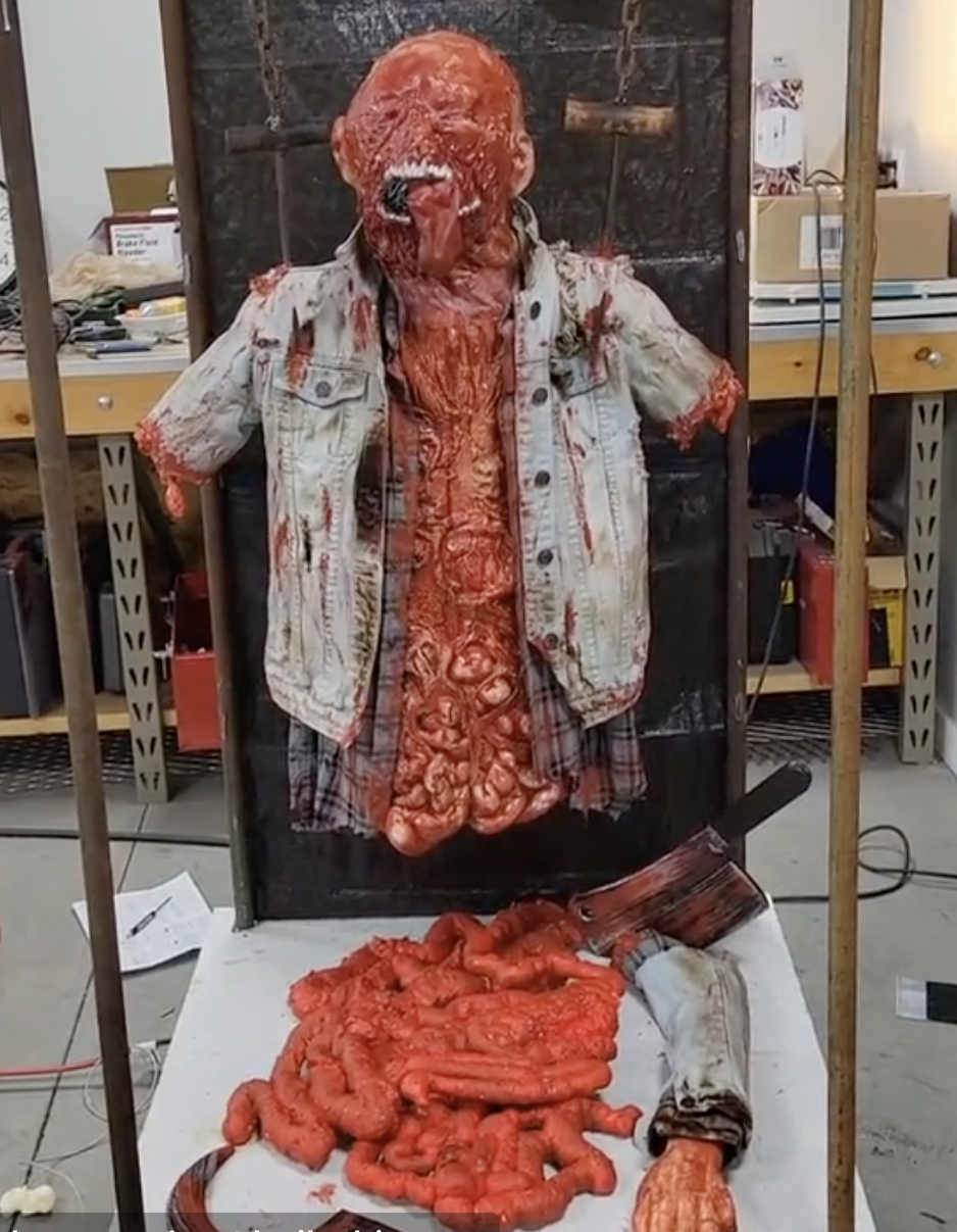 Horror prop of a zombie torso wearing a denim vest and shirt, with exposed bloody intestines and detached arm at the base, displayed in a workshop