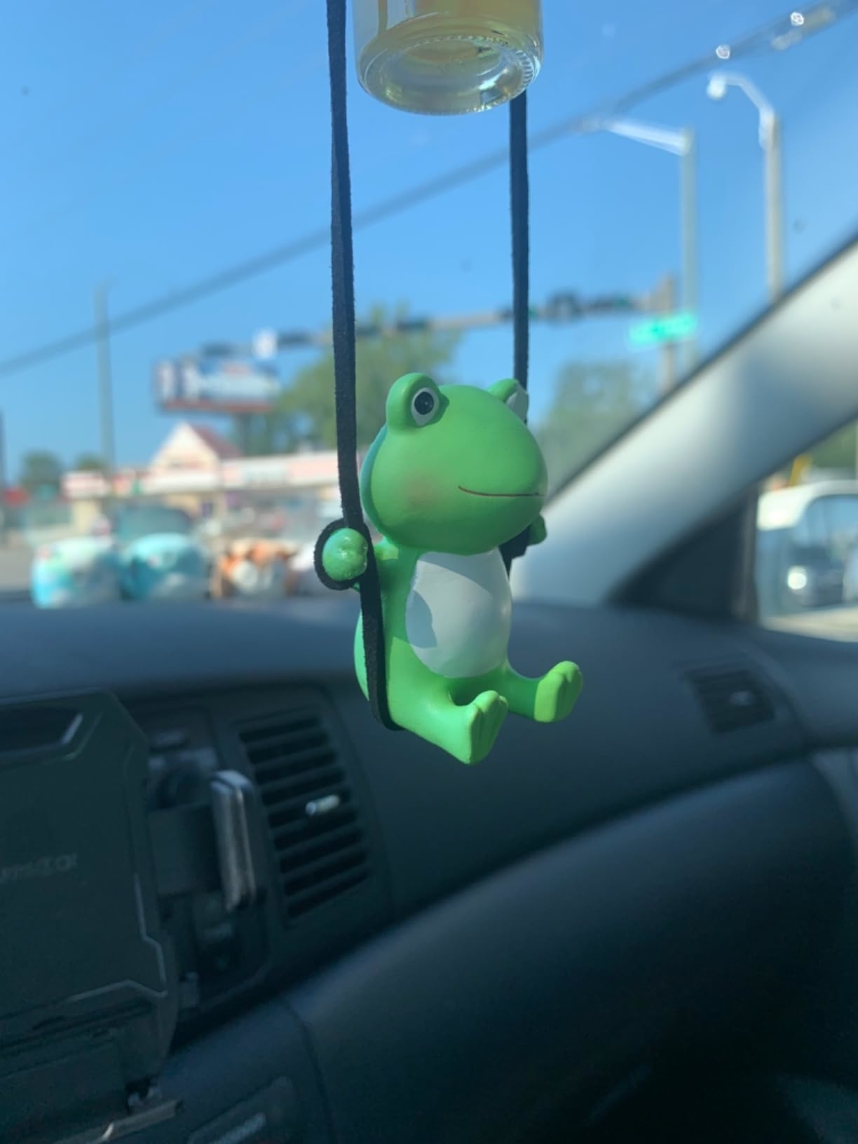 A green frog air freshener hangs from a car's rearview mirror, seen from the passenger seat inside a vehicle with a street outside