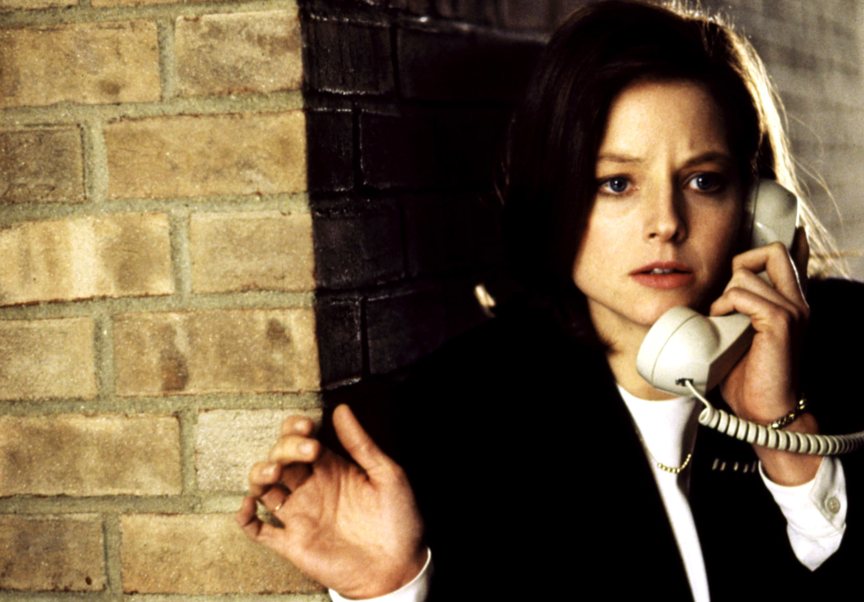 Jodie person is holding a phone, looking concerned, against a brick wall backdrop