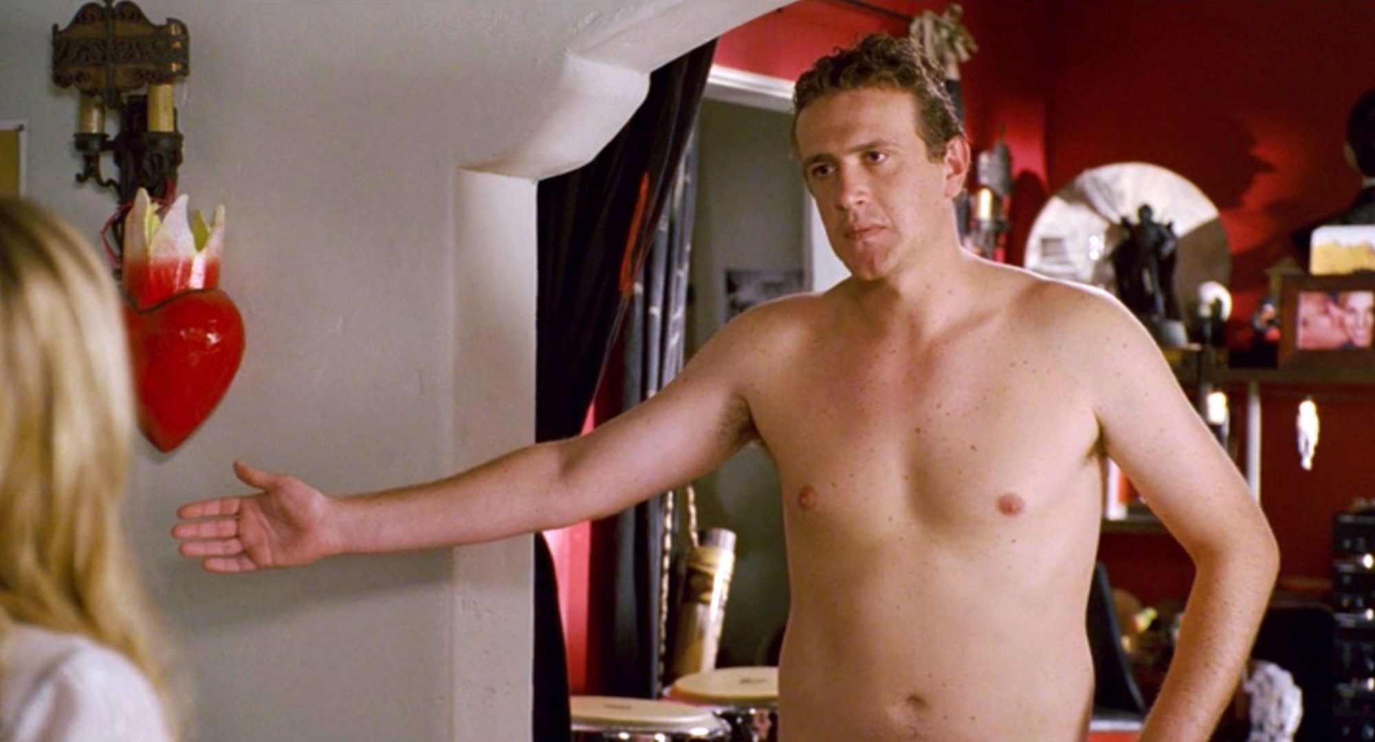 A shirtless man gesturing inside a home. Objects and art are visible in the background