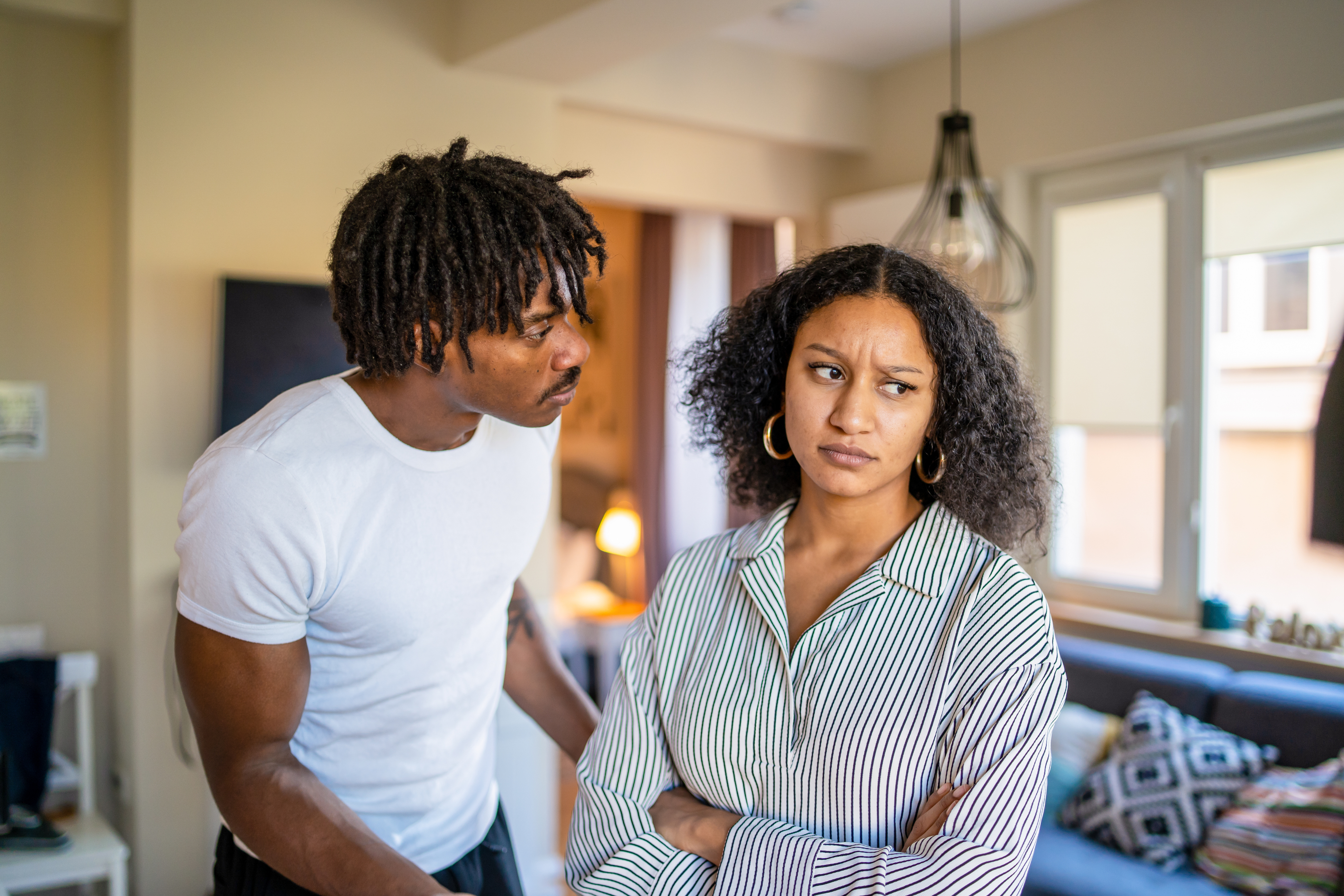 A couple at home appears to be in a tense conversation; the woman looks away with folded arms, while the man leans in towards her