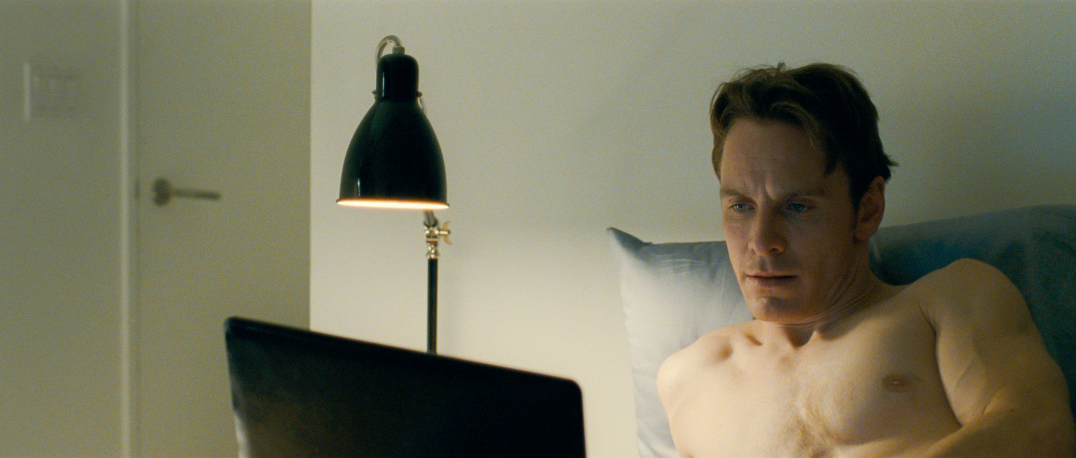 A shirtless man sits on a bed looking at a laptop in an indoor setting with minimal lighting