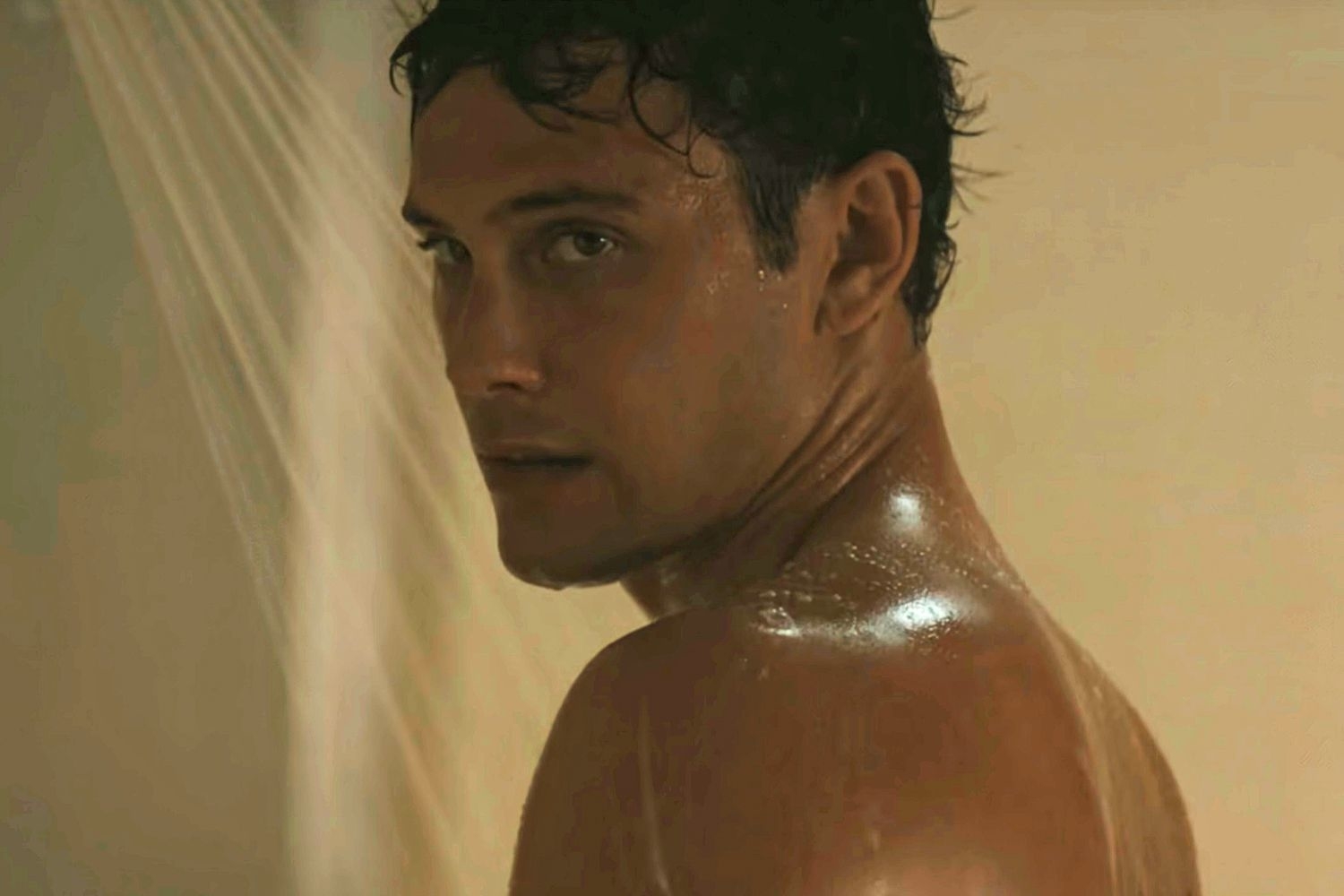 A shirtless person in a shower scene looks over their shoulder