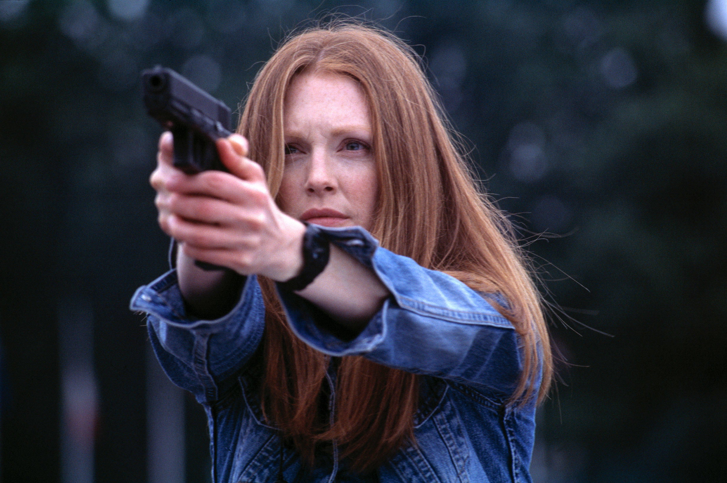 Julianne with long hair in a denim jacket points a gun forward. She looks focused and intense