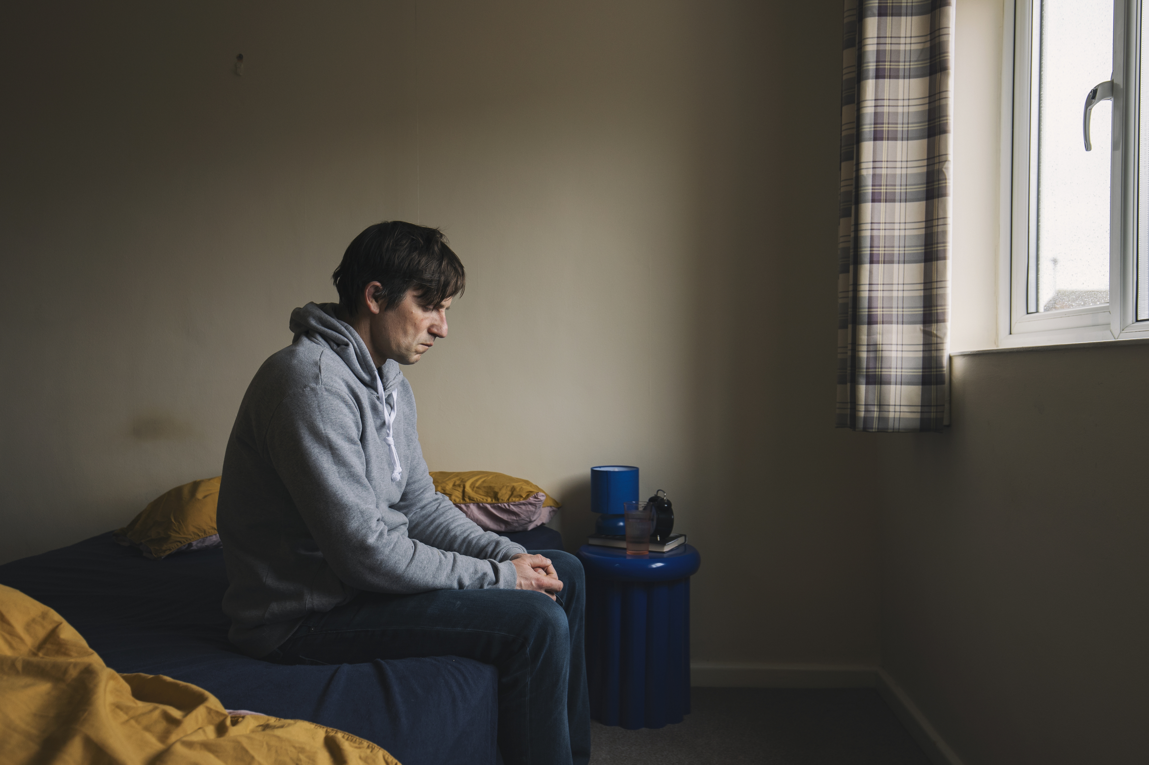 A person sitting on a bed in a pensive mood, facing a window. The setting is a simple, cozy room