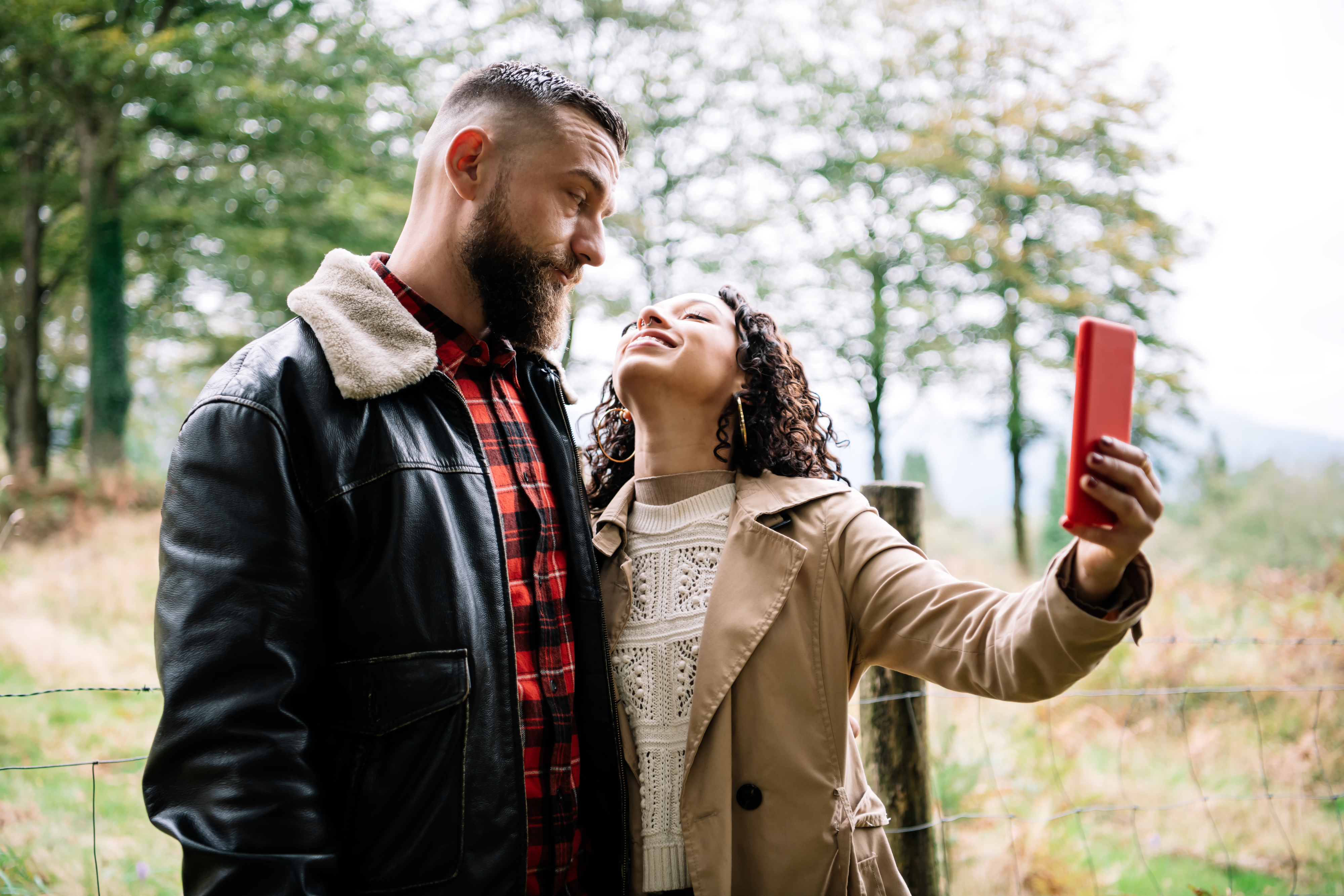 A couple takes a selfie outdoors, smiling at each other. One is wearing a leather jacket, and the other a beige coat