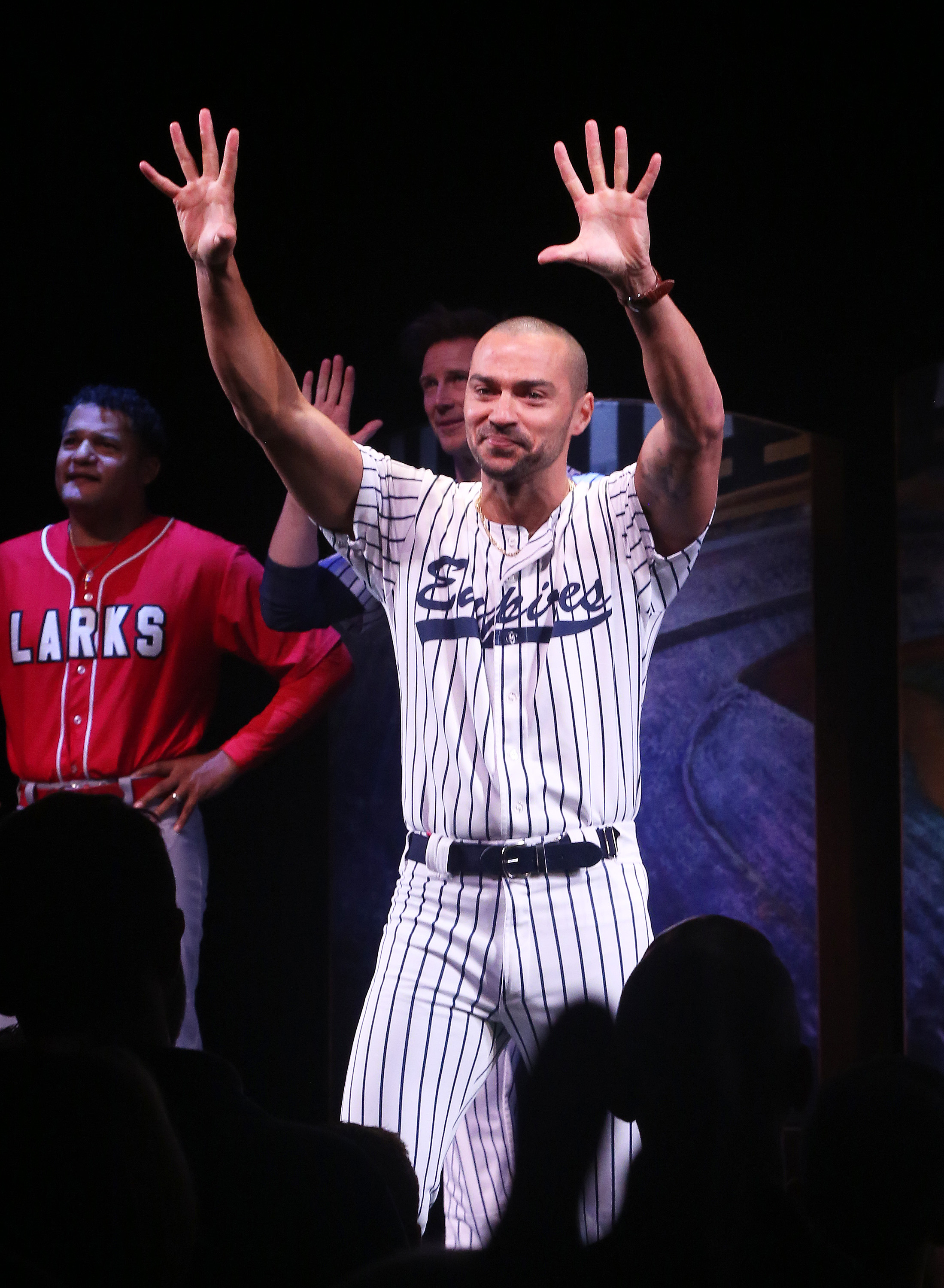 A man in a striped baseball uniform on stage, smiling and holding his hands up, with others behind him