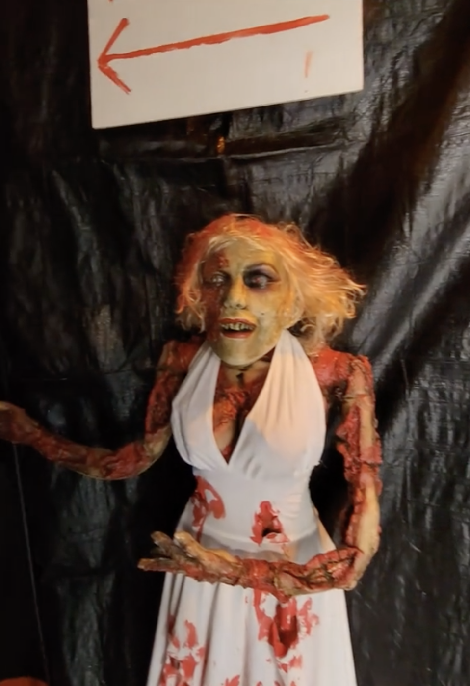 A zombie-like figure resembling Marilyn Monroe in a white dress with blood stains stands under a sign with an arrow