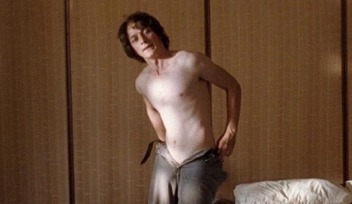 A shirtless man in a room, putting on pants, partially dressed