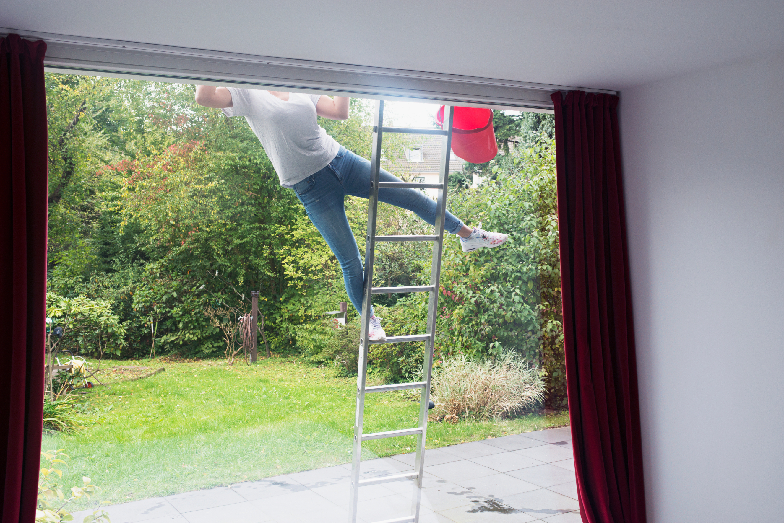 Person climbing a ladder by a large window, partially outside. Red curtains frame the view of a green garden