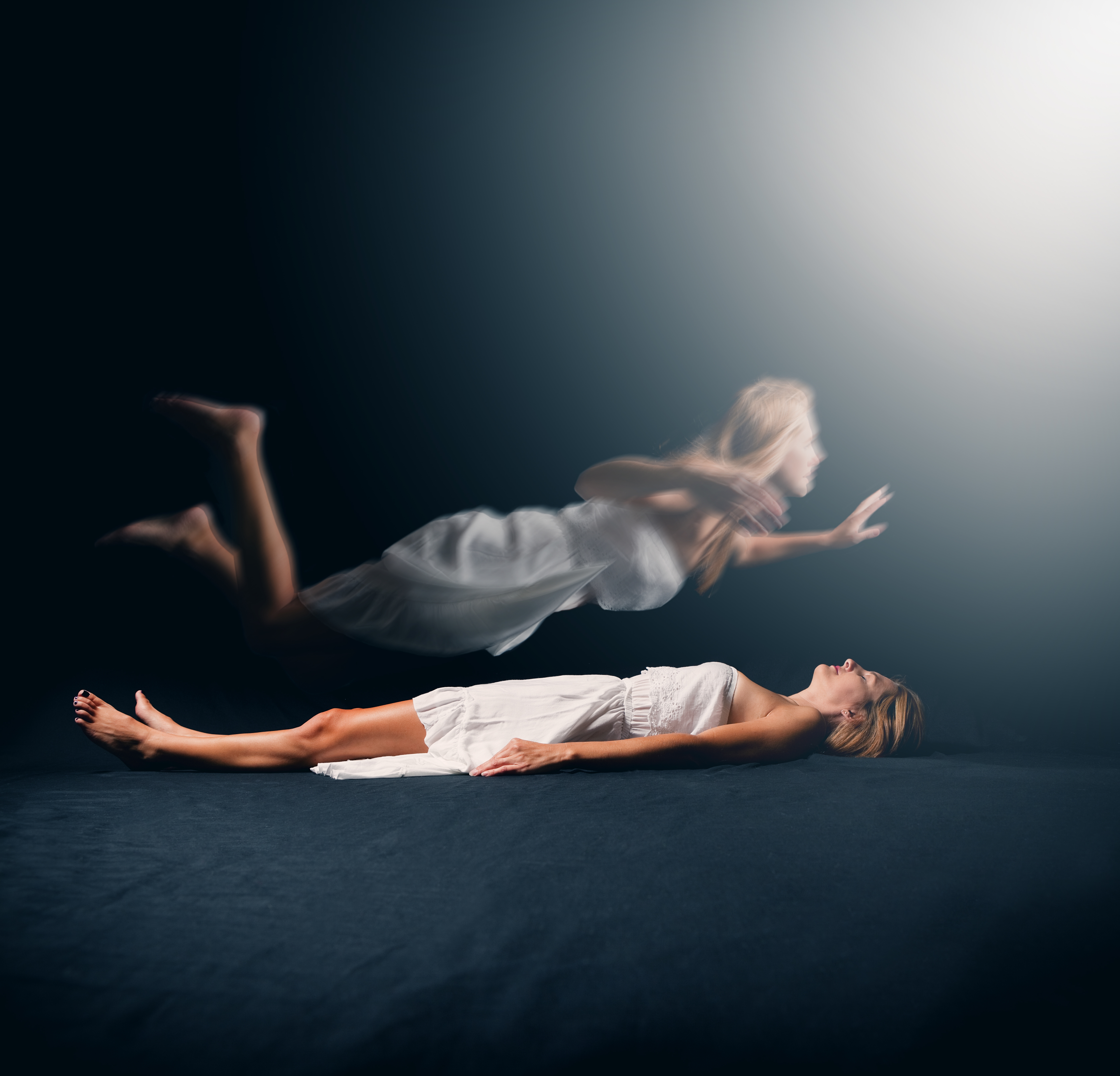 A woman lies on the ground in a white dress, with a ghostly version of herself rising above her